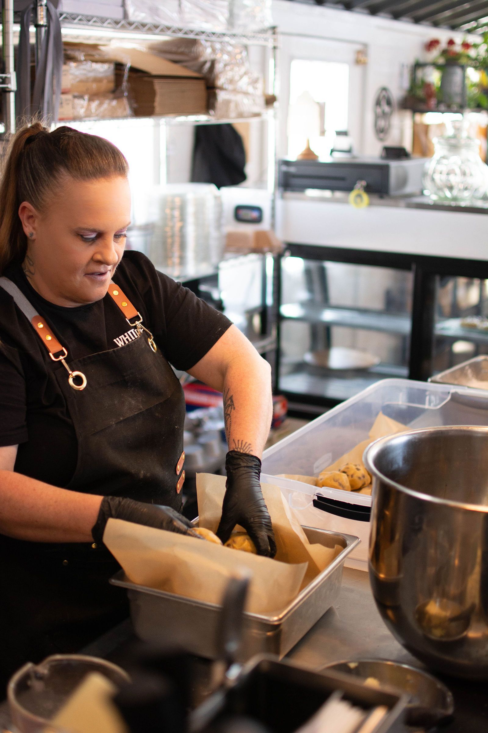 Person in black apron placing dough balls into a pan at a bakery.