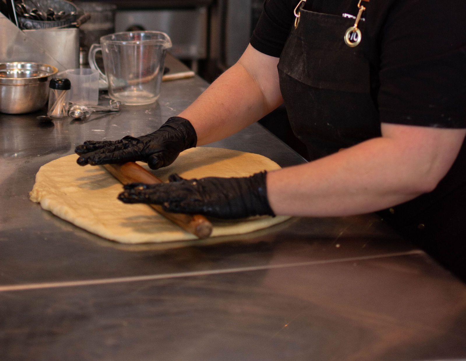 Person in black gloves rolling dough on a metal countertop.