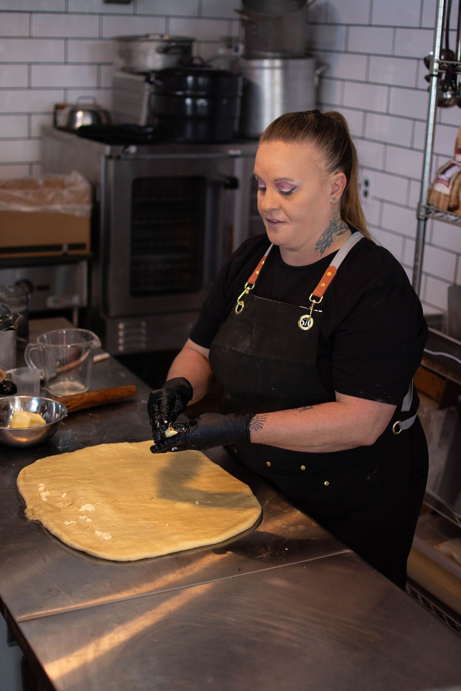 Woman in black apron spreads filling onto dough on a stainless steel surface in a kitchen.