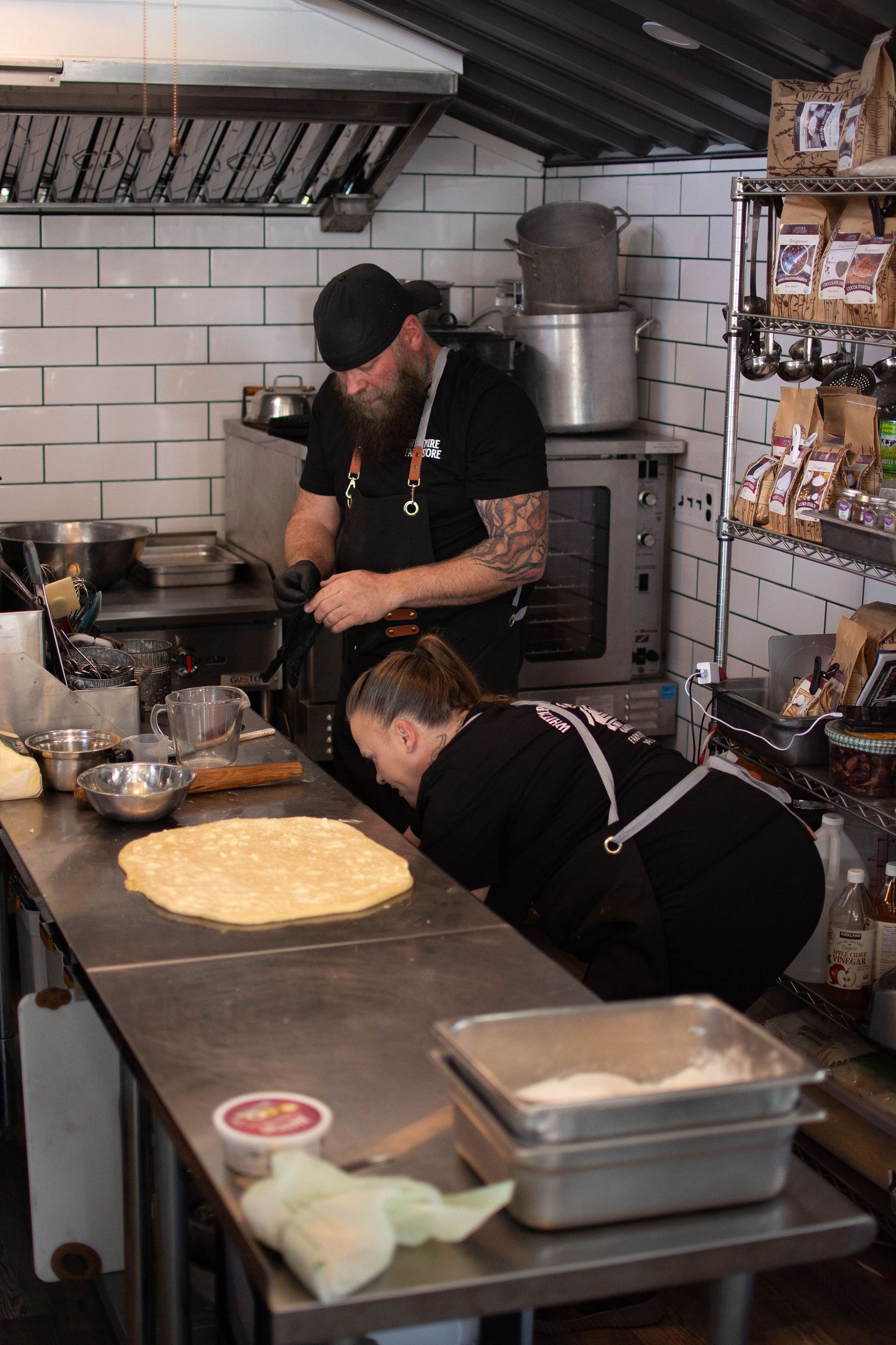 Two people in a commercial kitchen preparing food. One rolls dough, the other works by the stove.