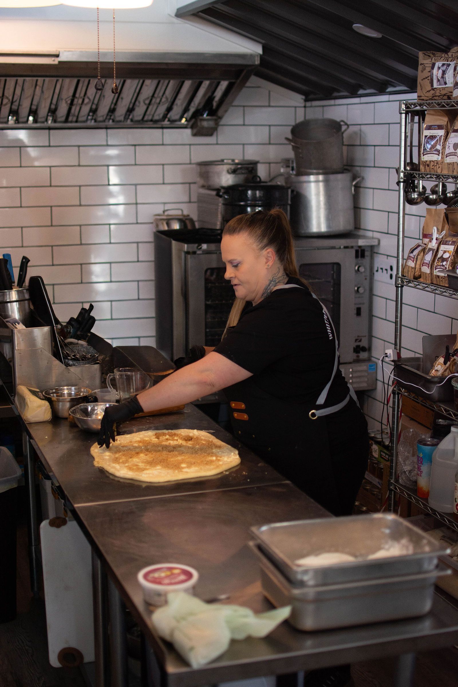 A person in a black apron spreads filling on a large, round pastry in a kitchen.