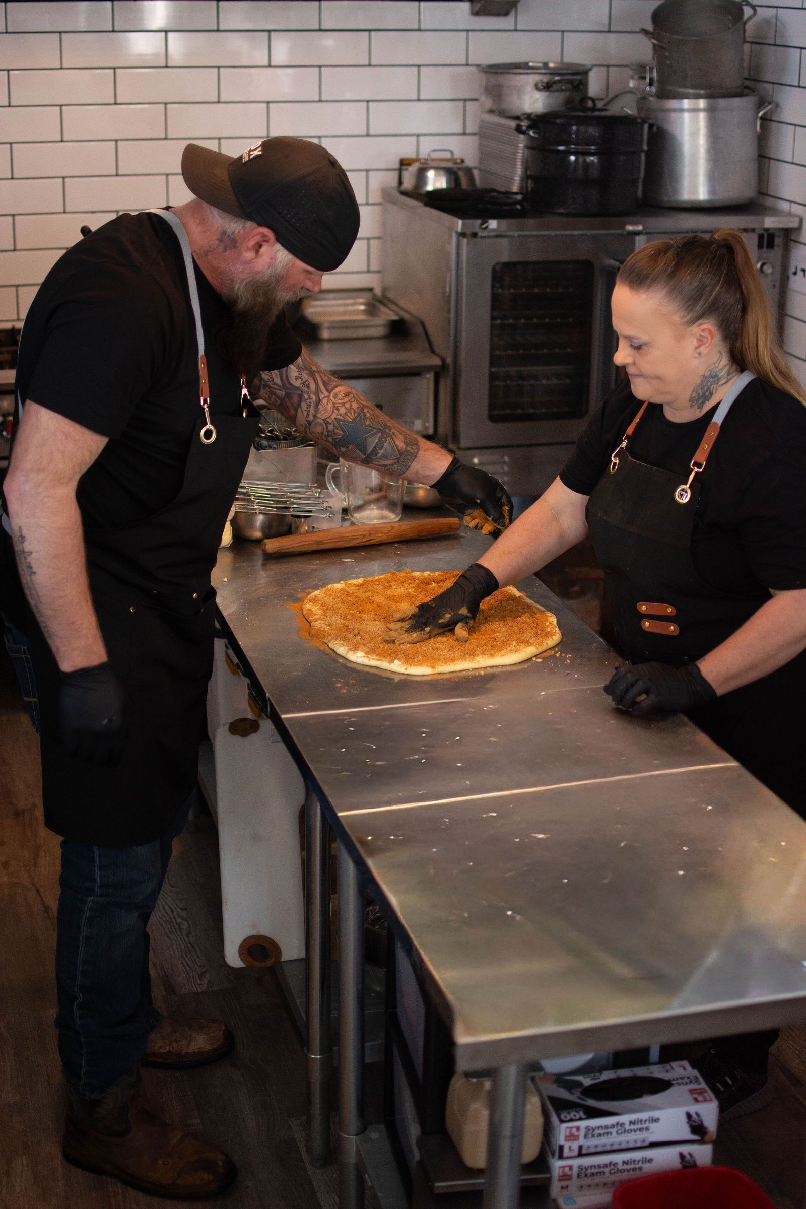 Two people in aprons and black gloves preparing food on a stainless steel countertop.
