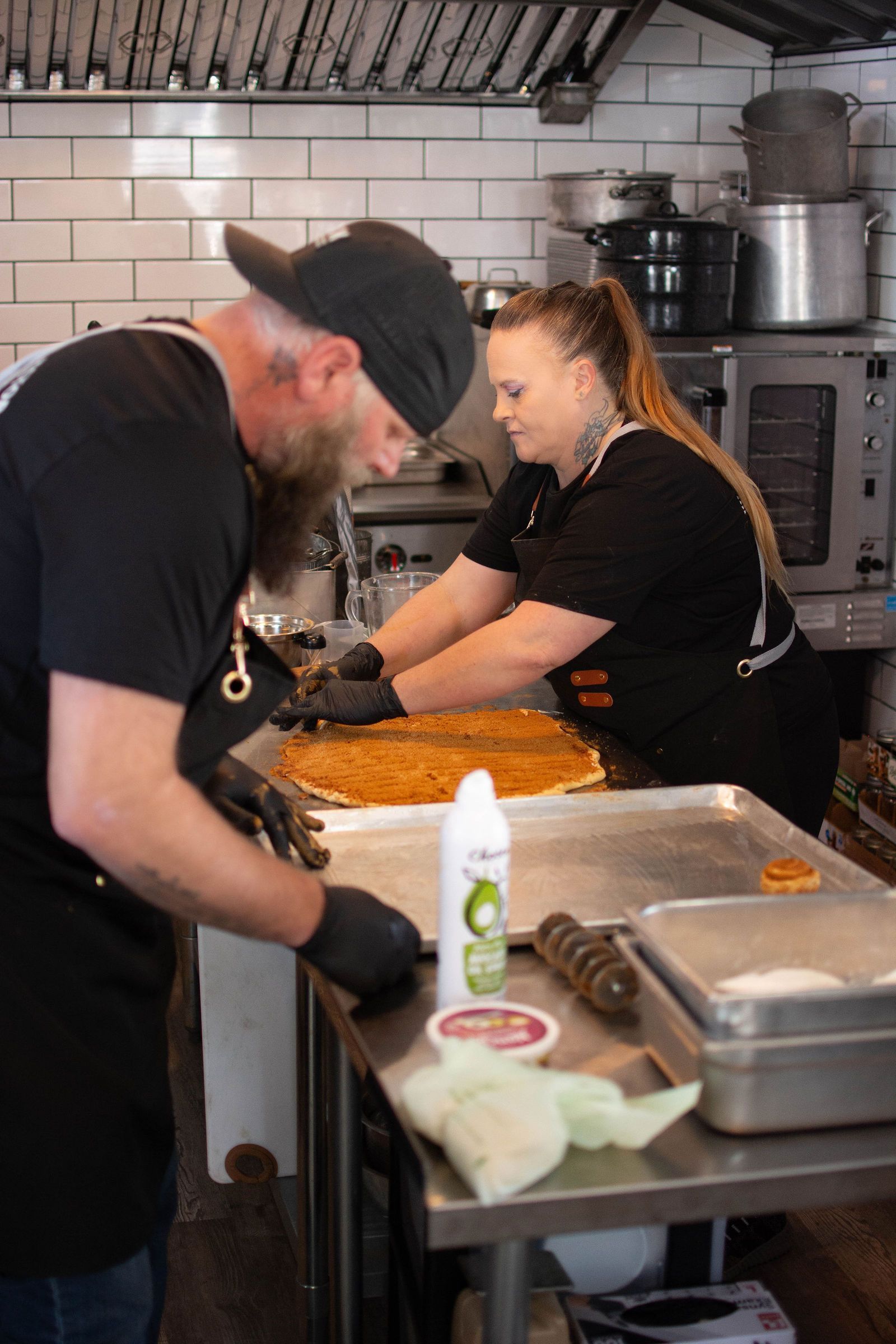 Two people preparing food in a commercial kitchen. One with a beard, in a cap; the other with a ponytail.