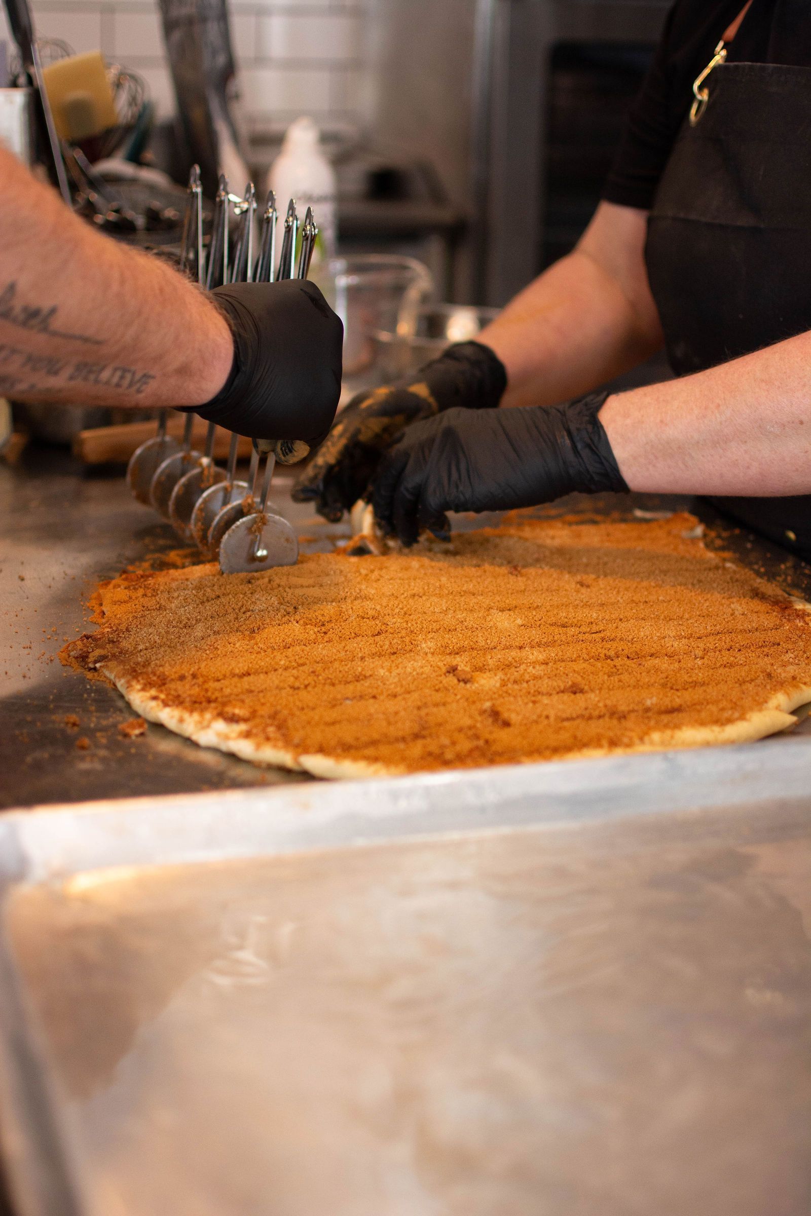 Two gloved hands cutting a large, circular, brown-dusted pizza on a metal countertop in a kitchen.