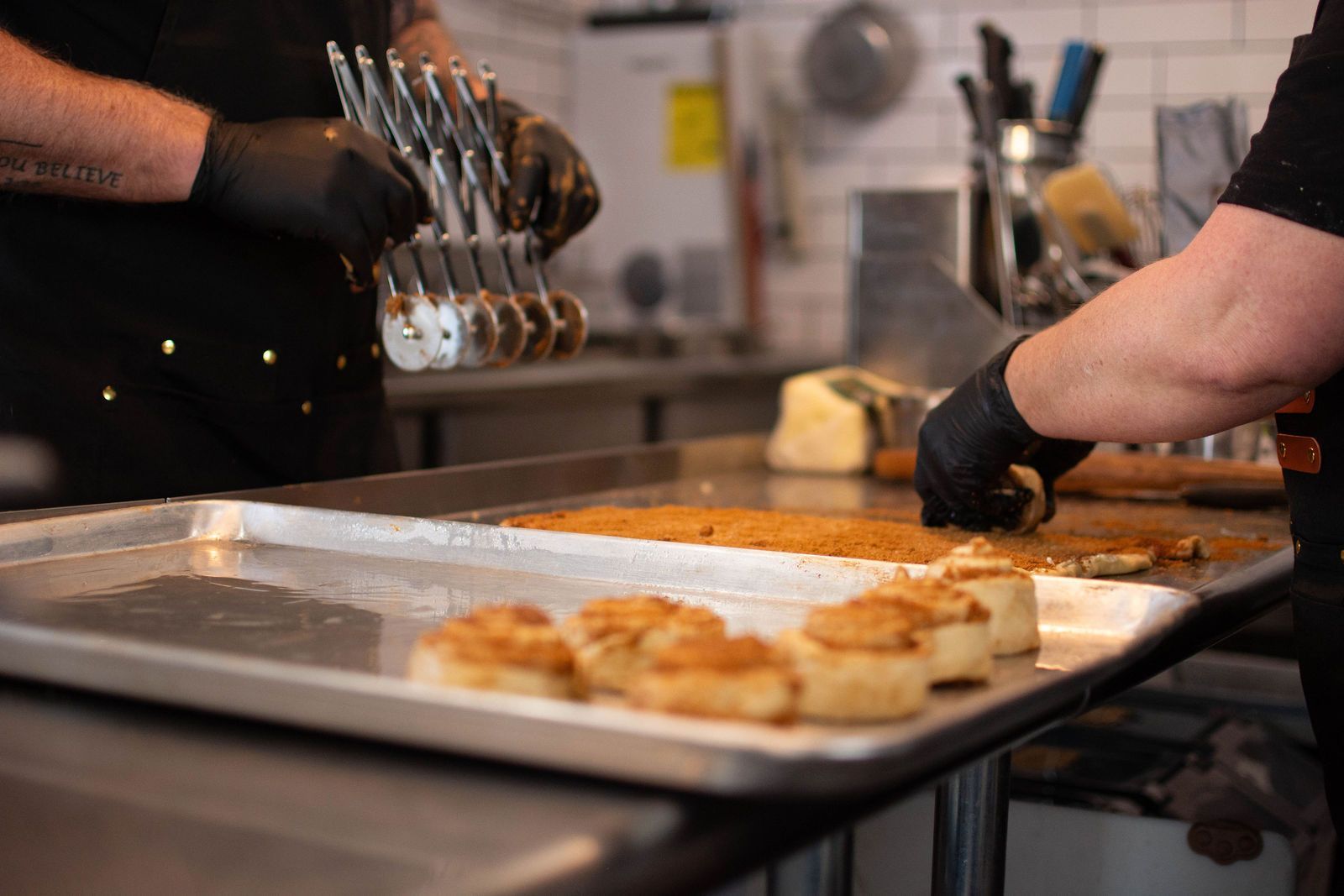 Two people in black gloves preparing food in a kitchen. A tray of baked goods is in the foreground.