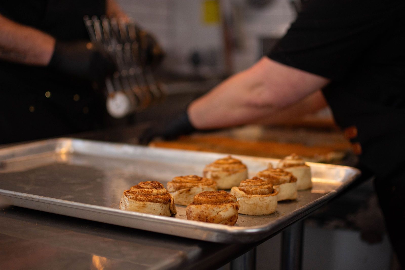 Cinnamon rolls on a baking sheet, person in black shirt and gloves preparing.