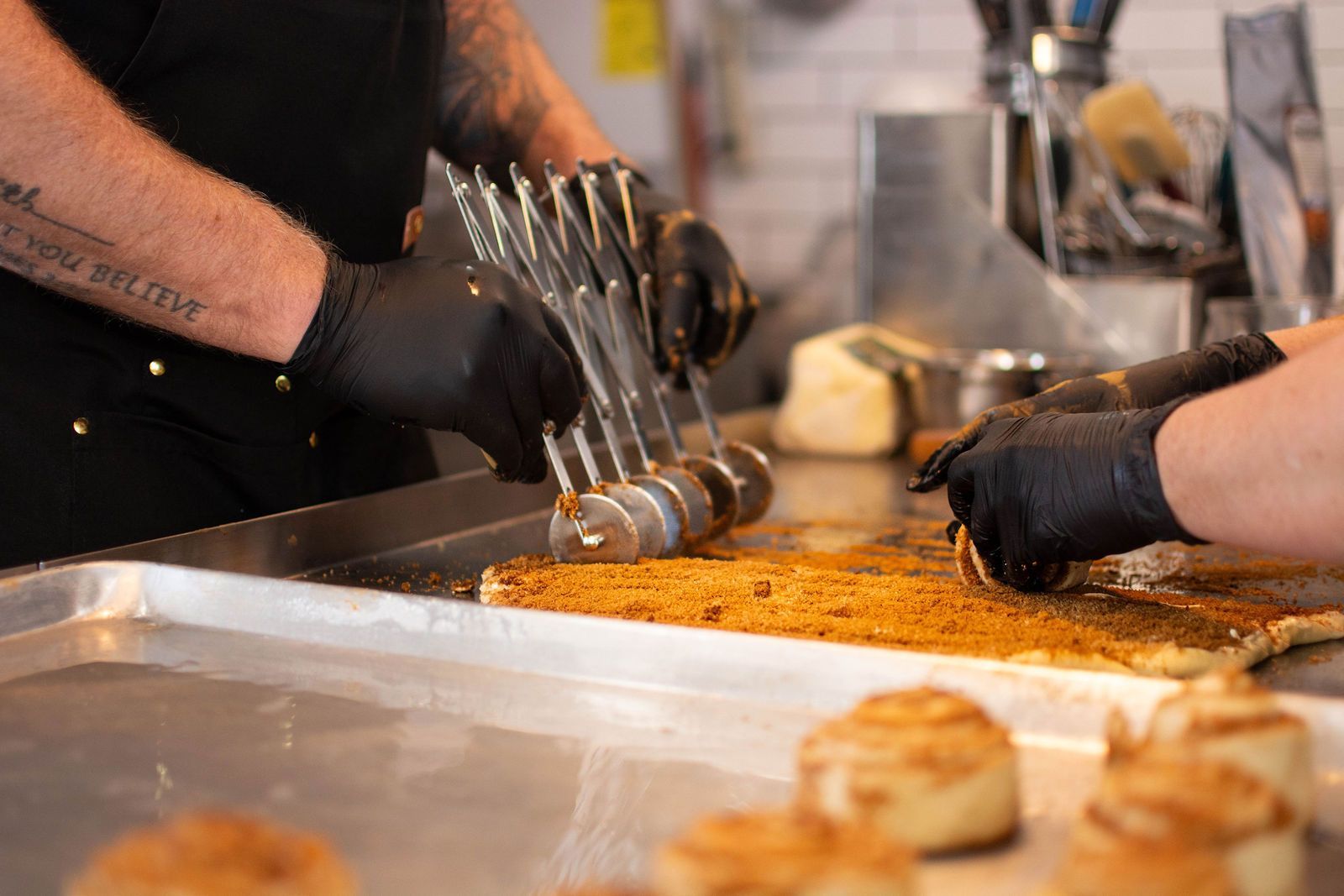 Hands in black gloves cutting cinnamon rolls on a metal tray with a multi-blade cutter.