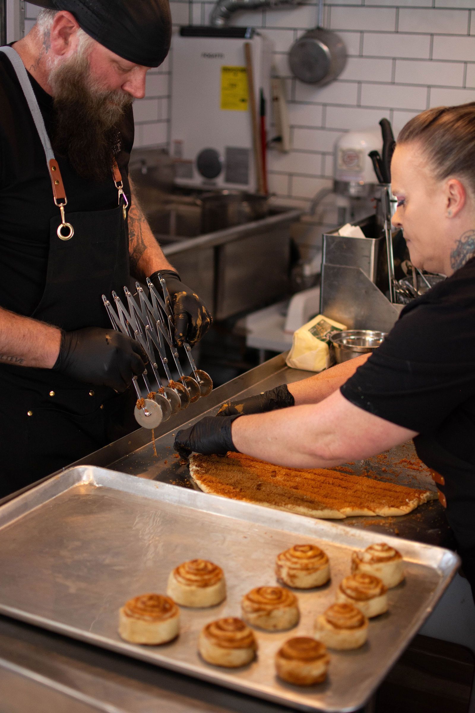 Two chefs preparing food in a kitchen. One cuts a pastry while the other holds metal skewers. Cinnamon rolls in foreground.