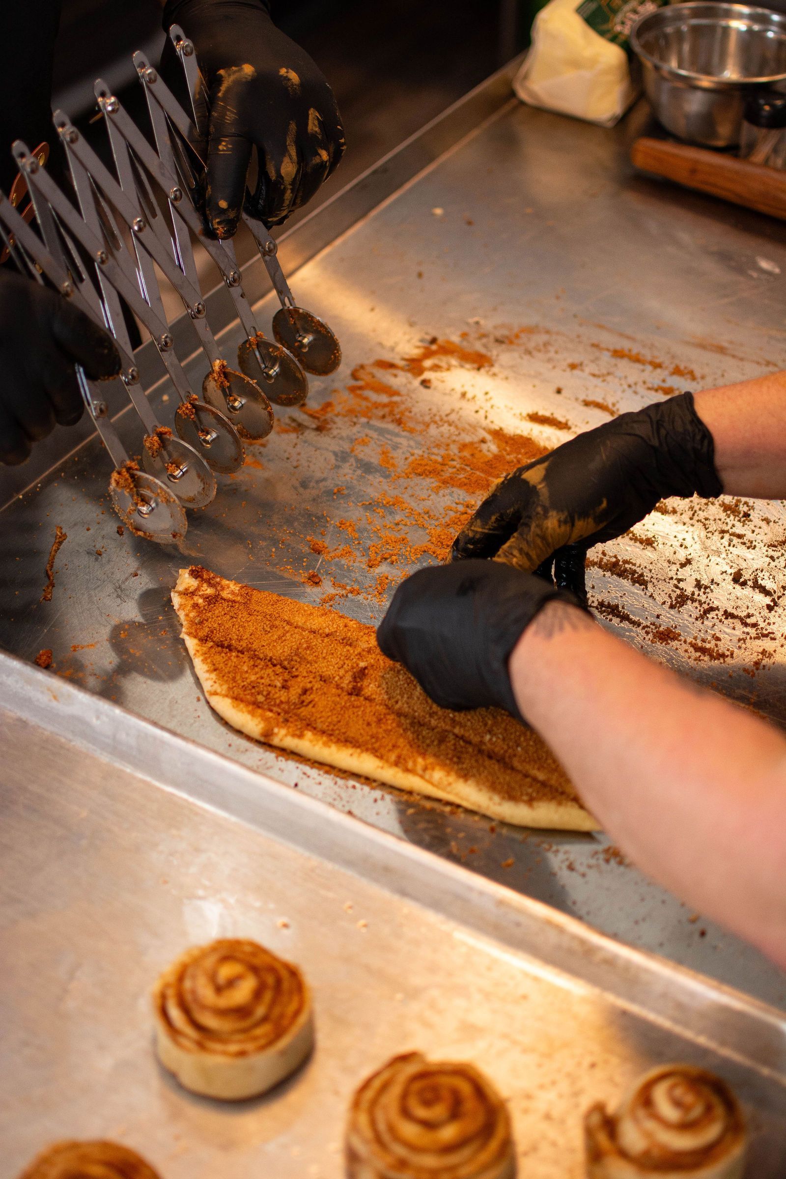 A person cuts cinnamon rolls, covered in cinnamon, with a multi-blade cutter on a metal counter.