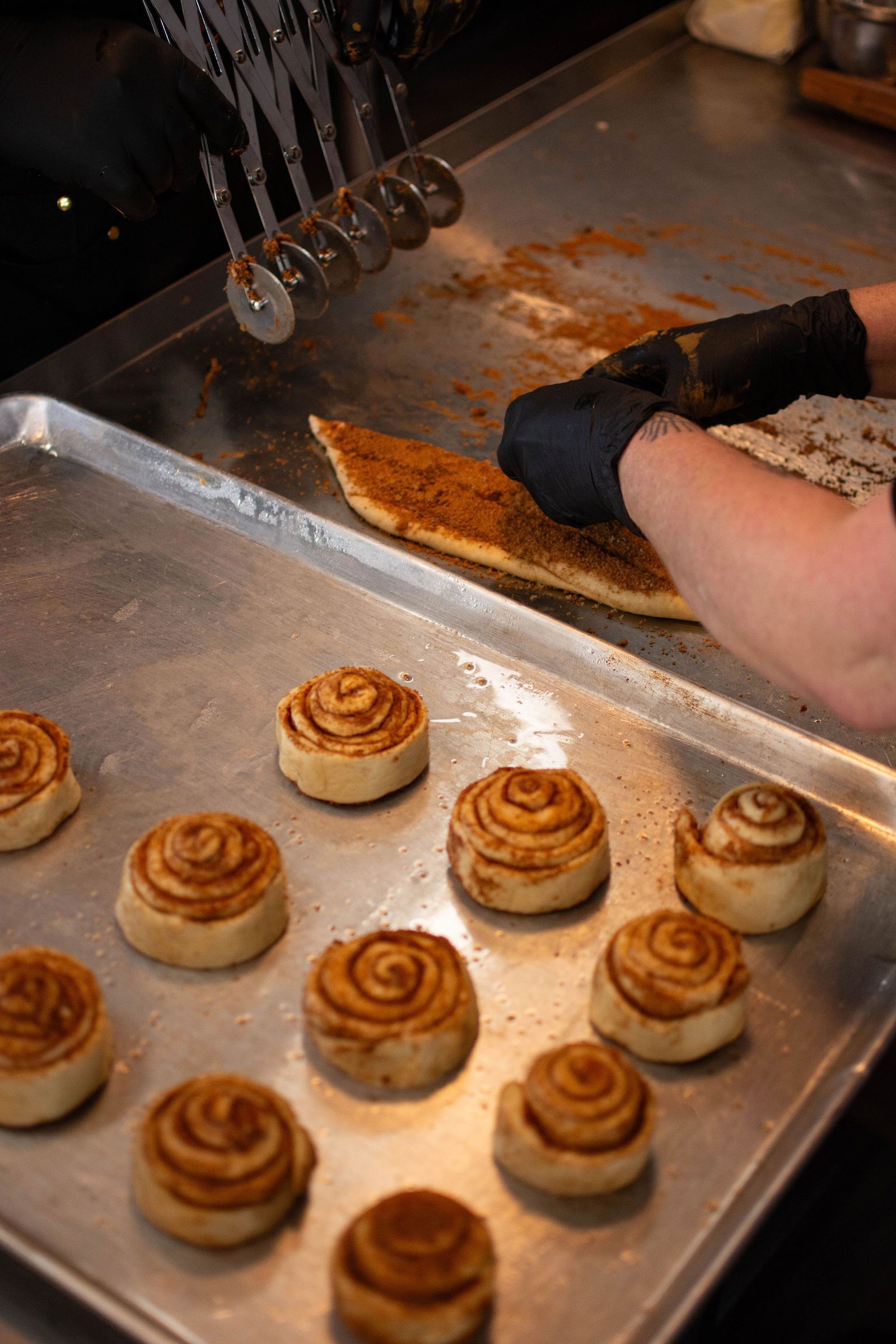 Baker cuts cinnamon roll dough on a metal tray, placing rolls onto a baking sheet.