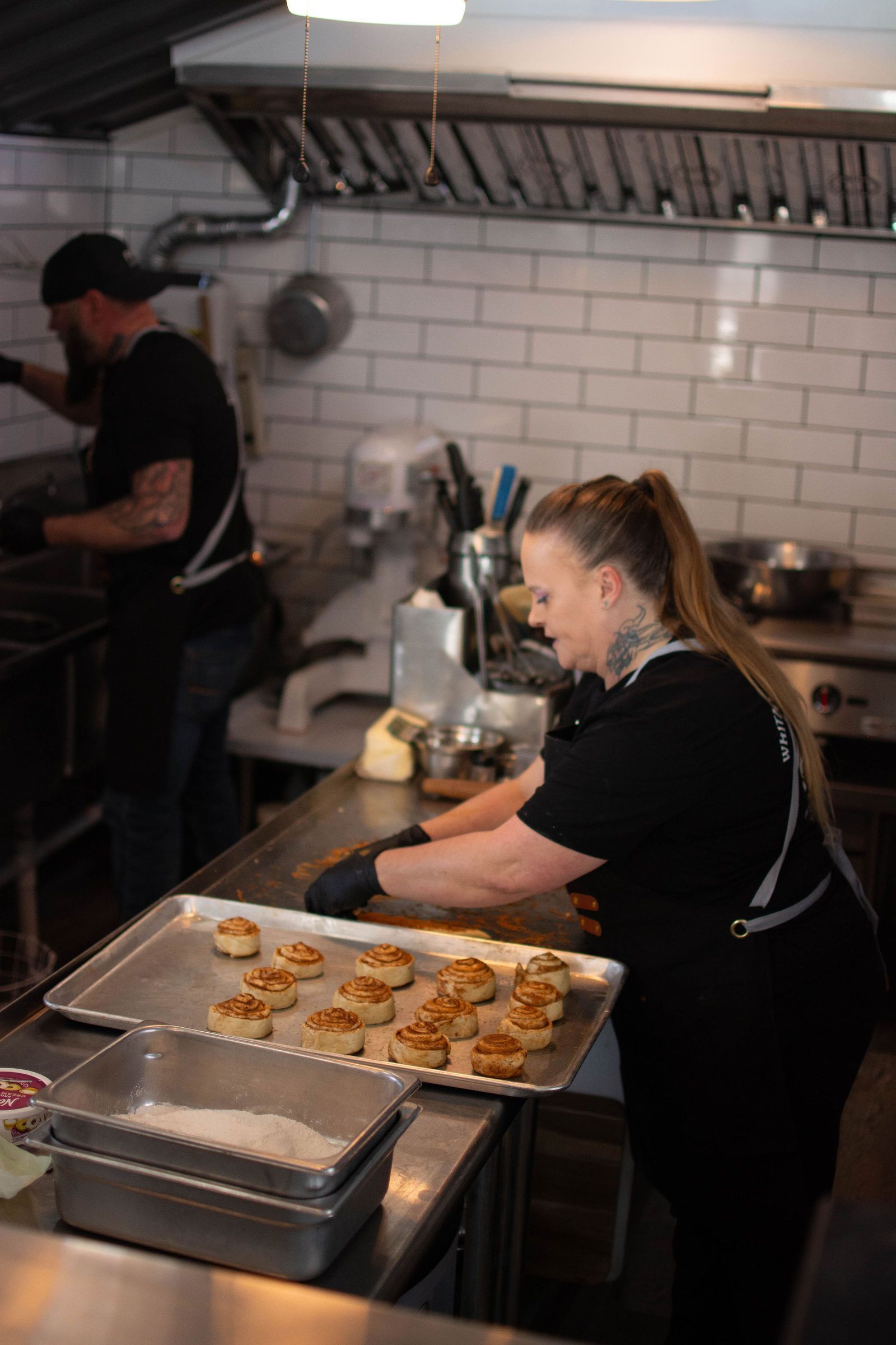 Two people in a commercial kitchen; one places tray of pastries, other at stove.
