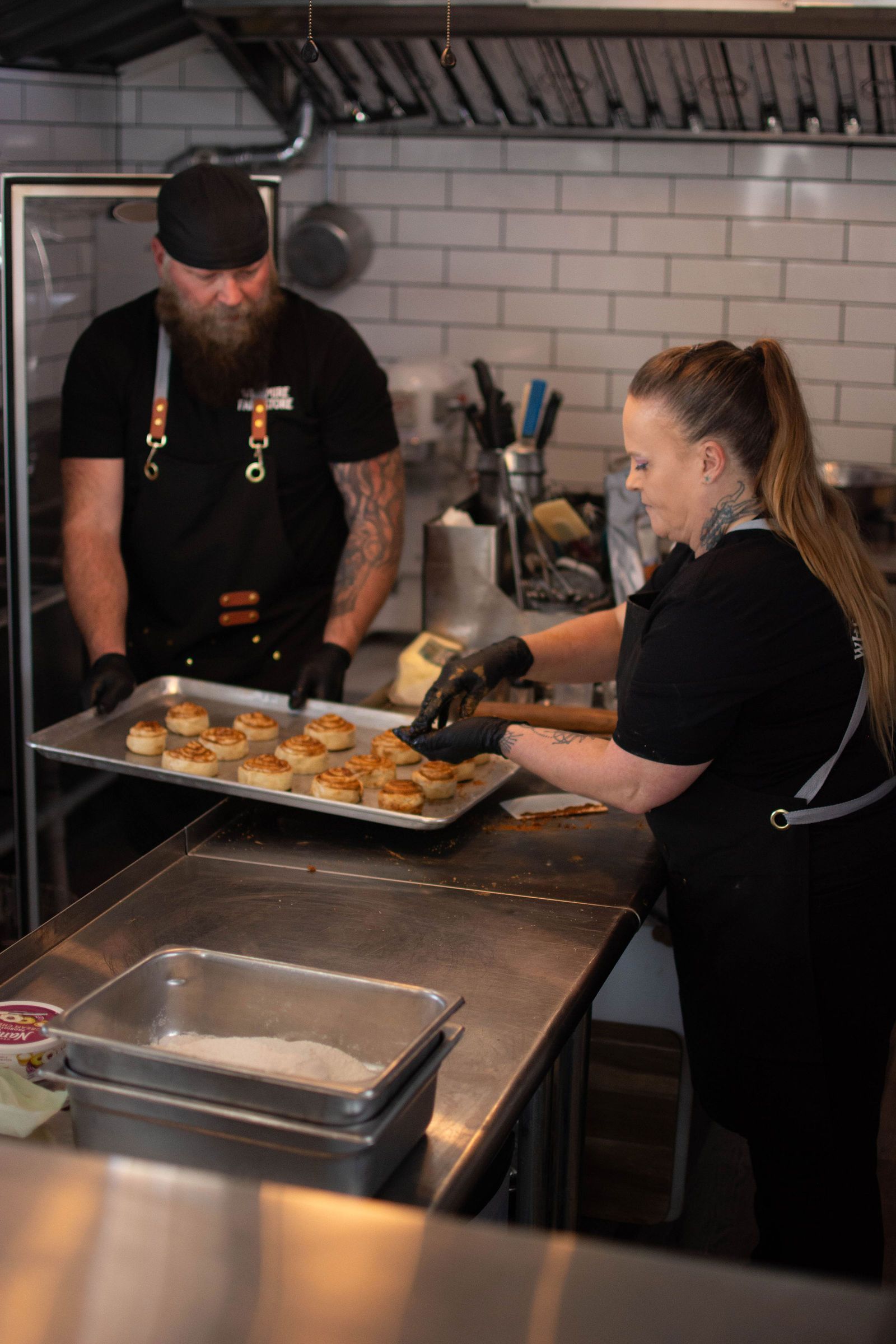 Two people in a kitchen preparing food, one holding a tray of buns.
