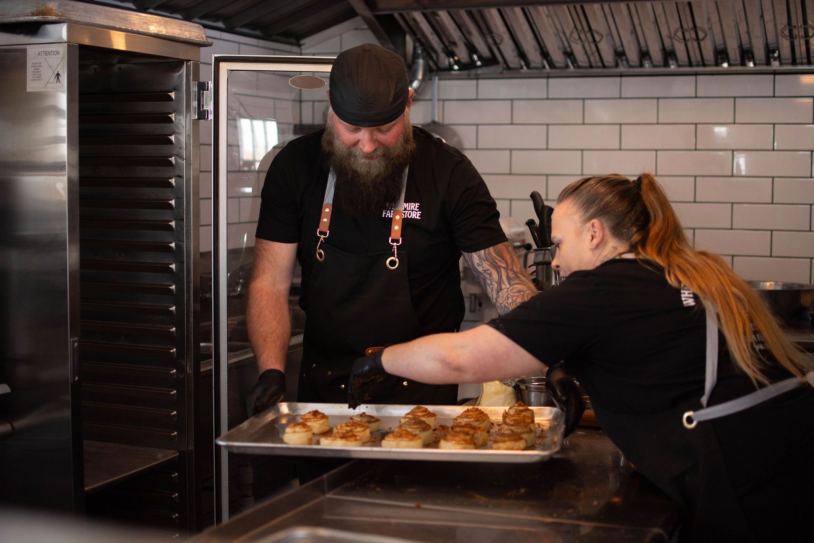 Two cooks in a commercial kitchen preparing food. One loads a tray of food into an oven.