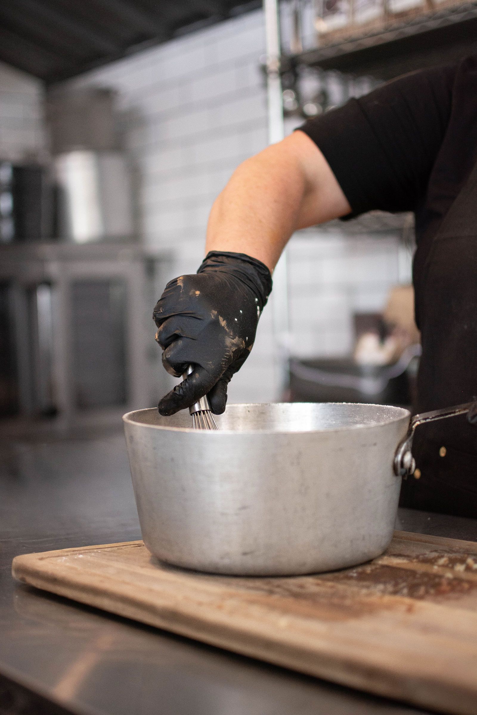 Person wearing a black glove stirring a pot with a handle on a wooden cutting board in a kitchen.