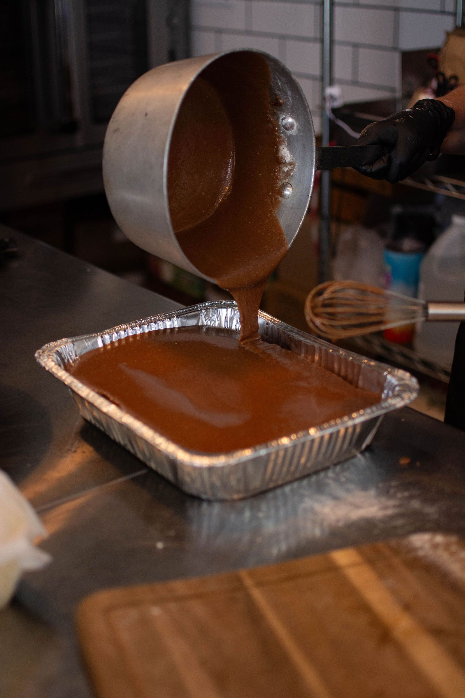 Brown batter being poured from a metal pot into a foil baking pan on a stainless steel surface.