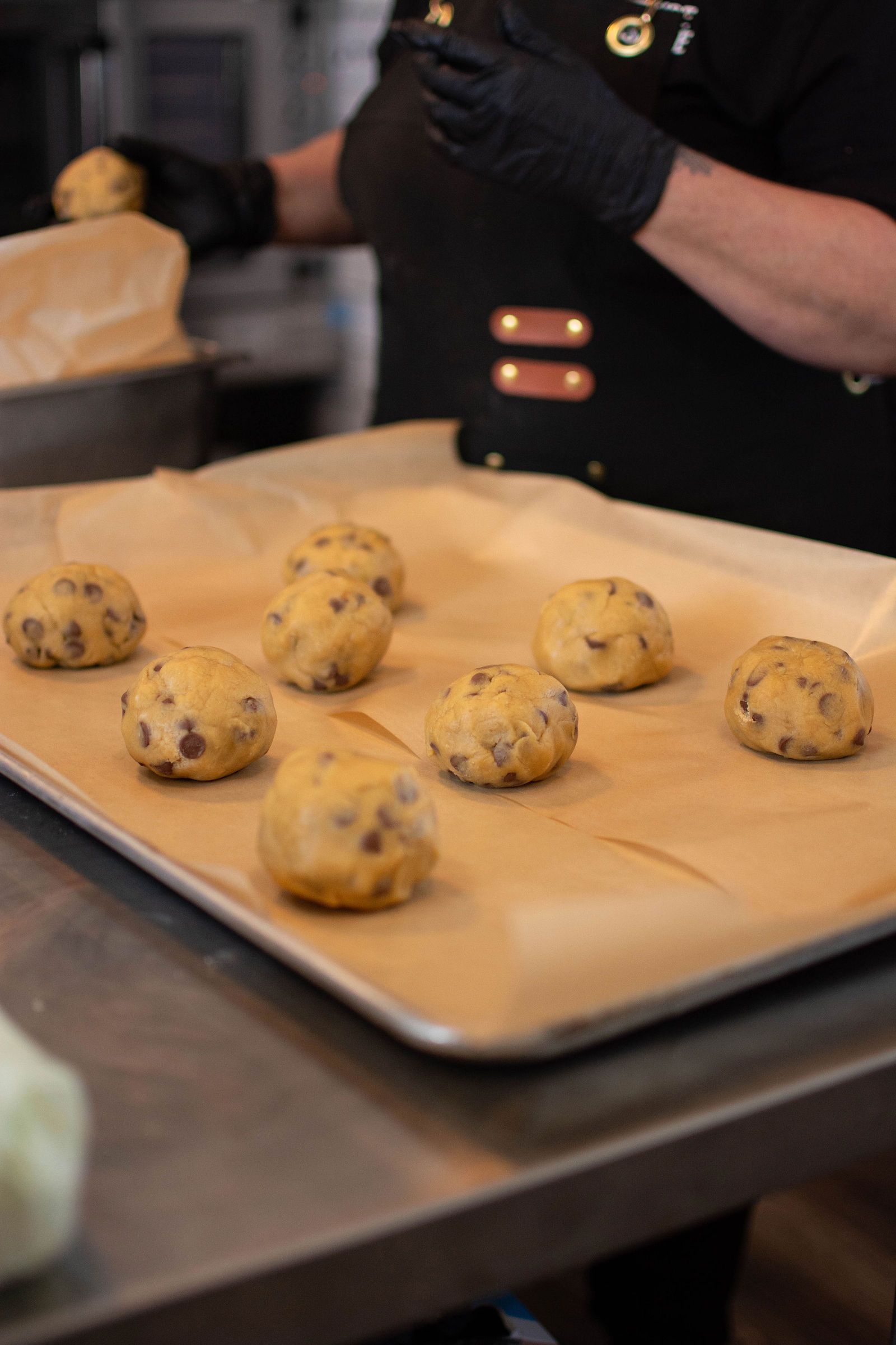 Person wearing black gloves placing cookie dough balls on a baking sheet lined with parchment paper.