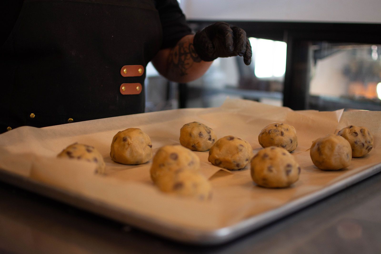 Cookie dough balls on a baking sheet, person in black gloves reaching above.