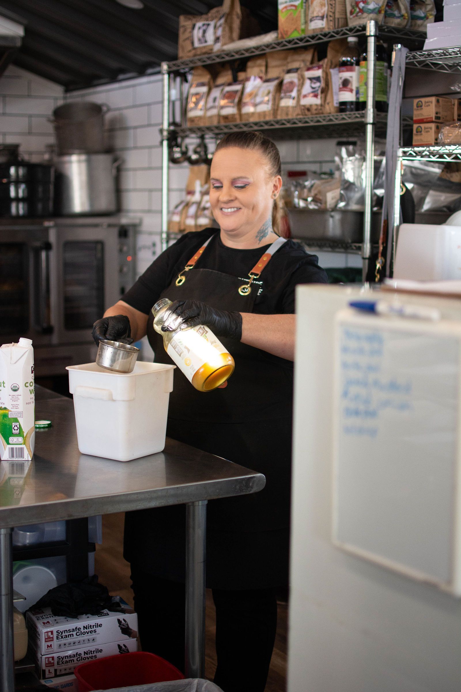 Woman in apron pouring liquid from a bottle into a container at a stainless steel counter.