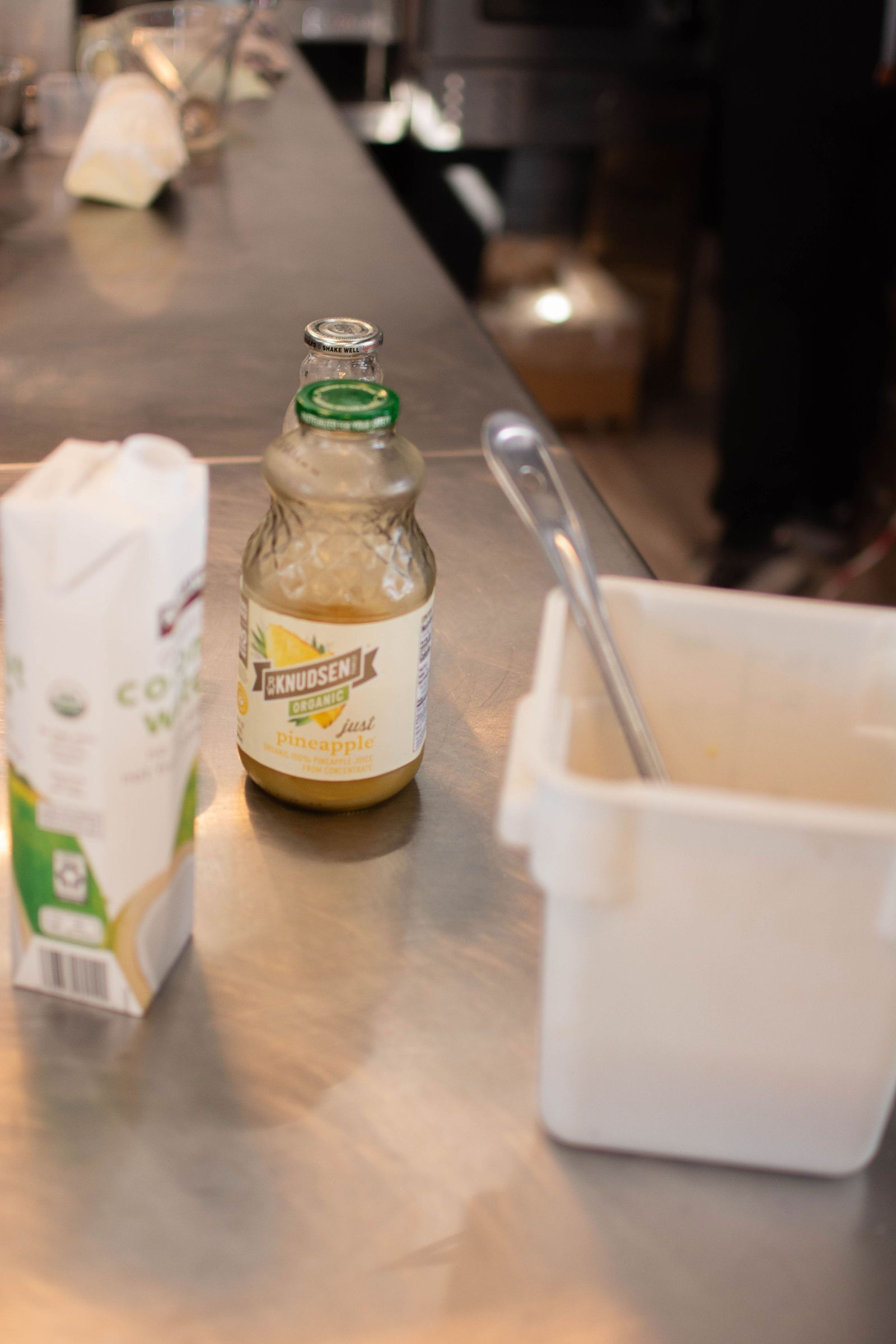 A bottle of lemonade, a container with a spoon, and a bag of flour sit on a stainless steel counter.
