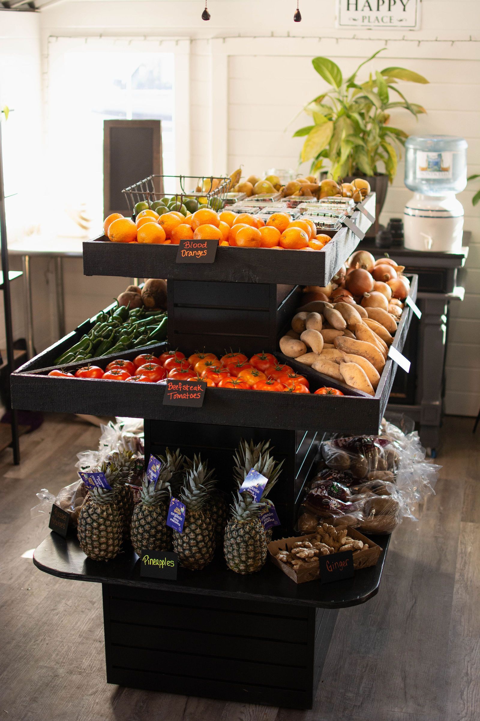A multi-tiered display of produce, including pineapples, tomatoes, and oranges, inside a brightly lit store.