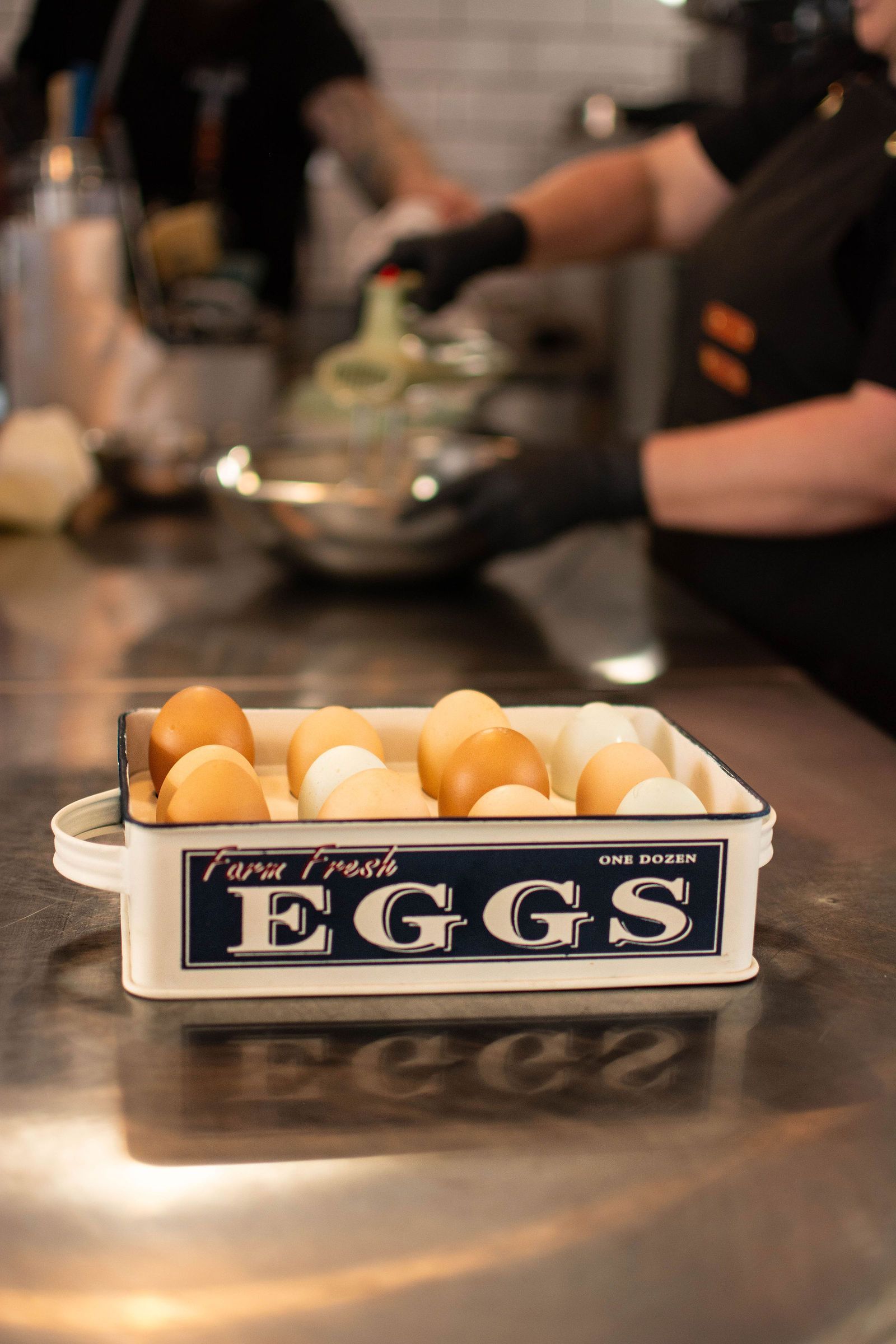Farm fresh eggs in a vintage ceramic carton on a stainless steel counter, chef in background.