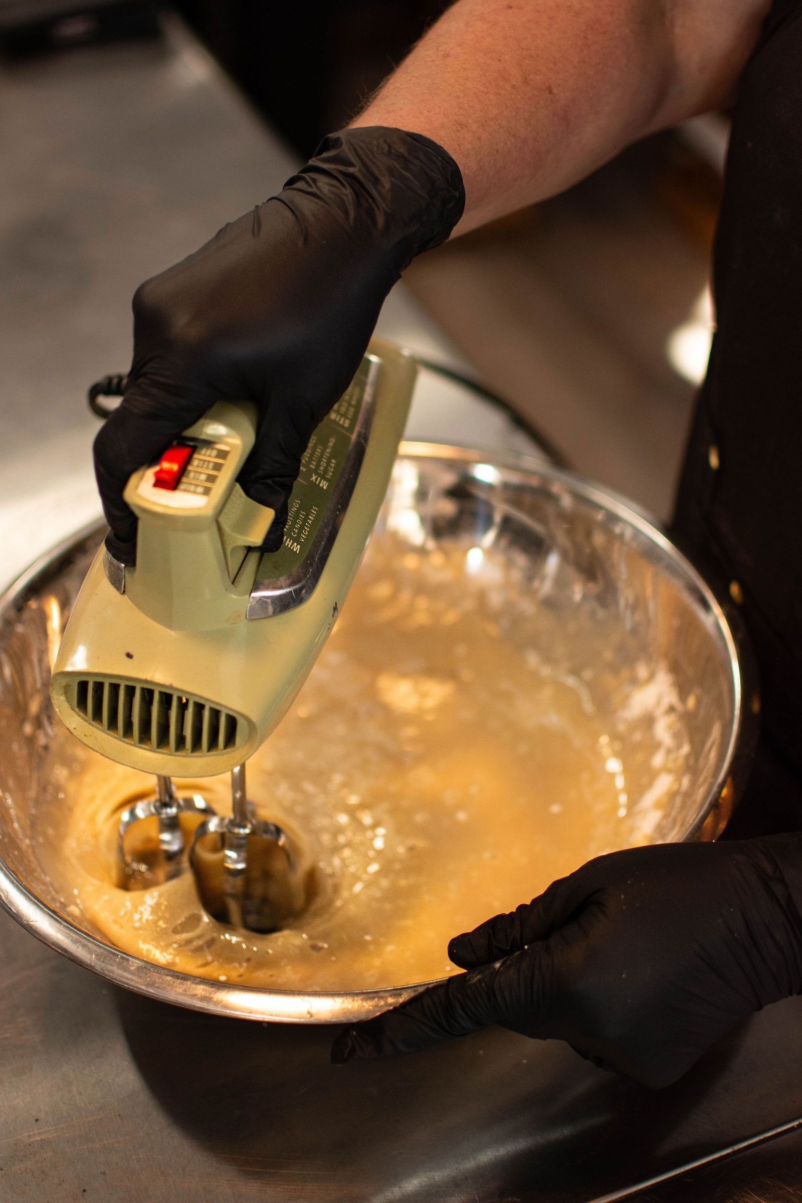A person in black gloves uses a hand mixer to blend ingredients in a stainless steel bowl.