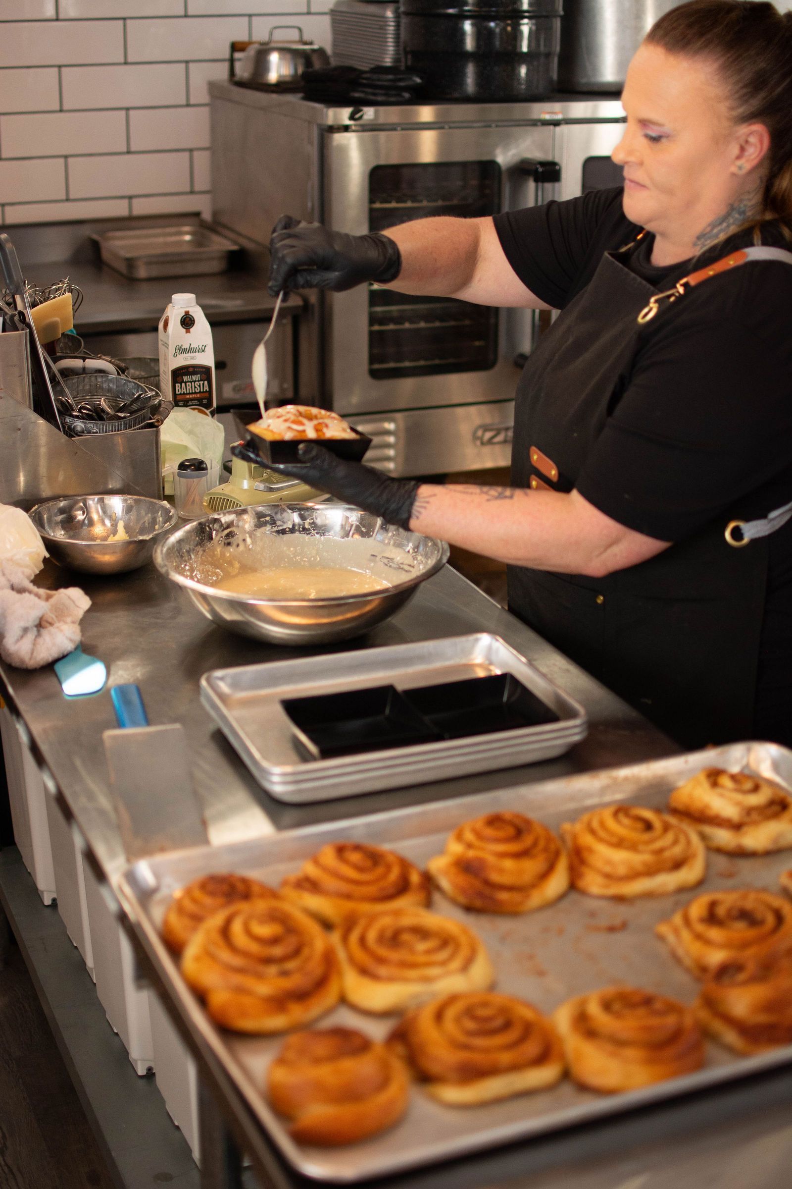 Chef in black apron glazes pastry in a commercial kitchen. Cinnamon rolls on a tray, ovens in background.