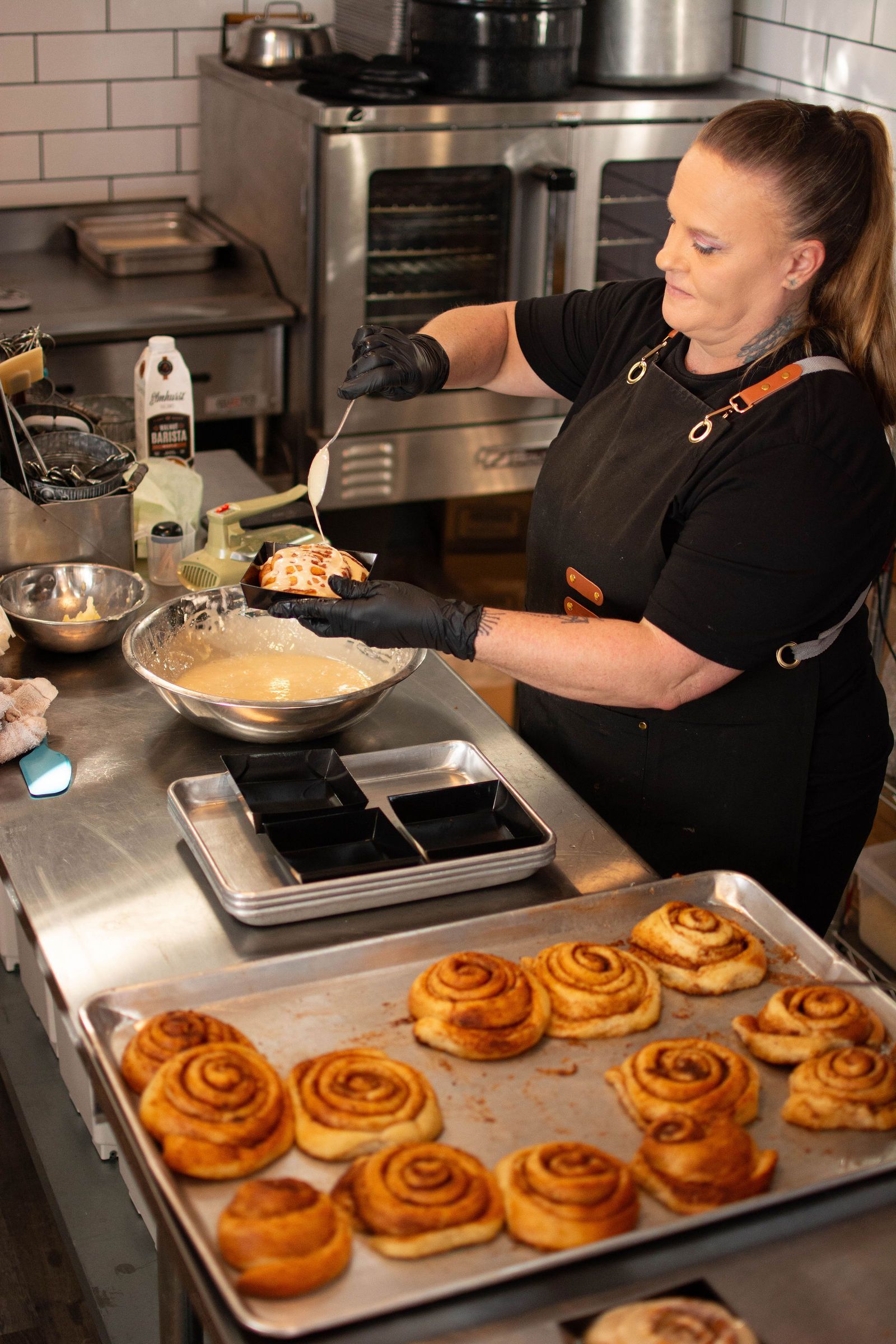Woman in black apron frosting cinnamon rolls in a kitchen, using black gloves.
