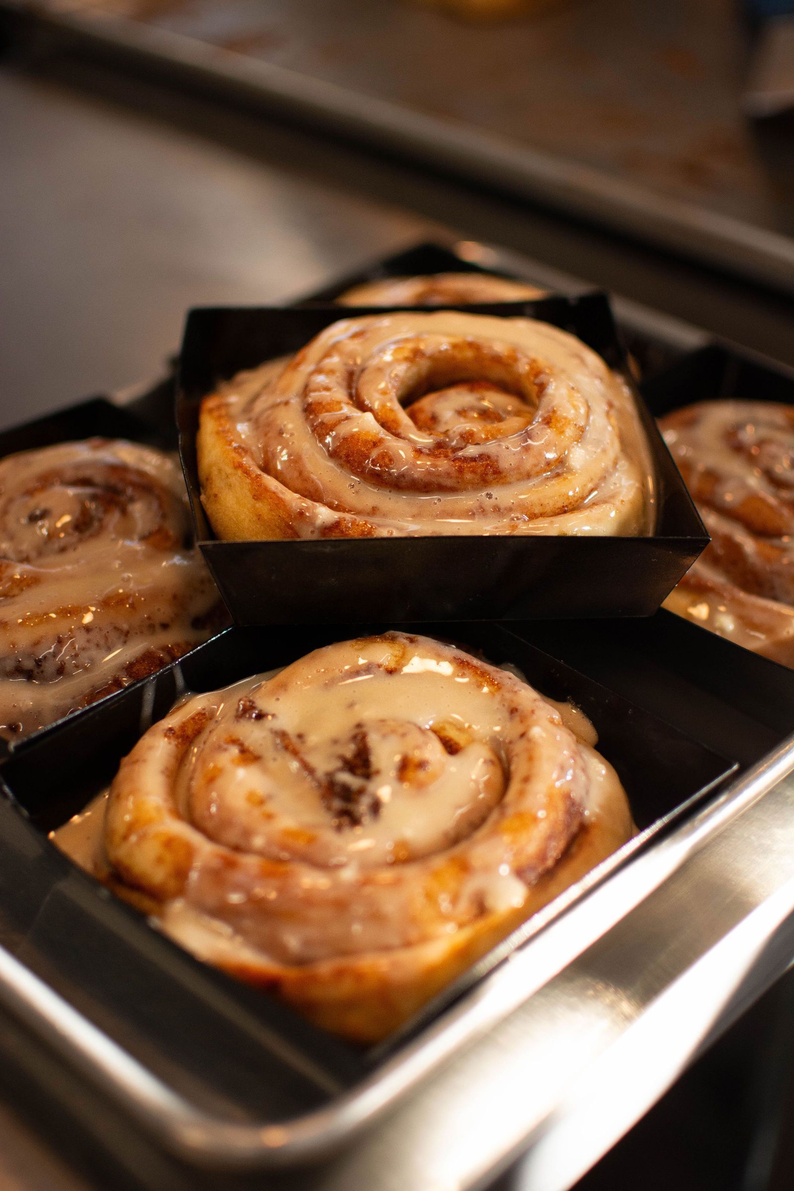 Cinnamon rolls with glaze in individual black paper trays, in a metal baking pan.