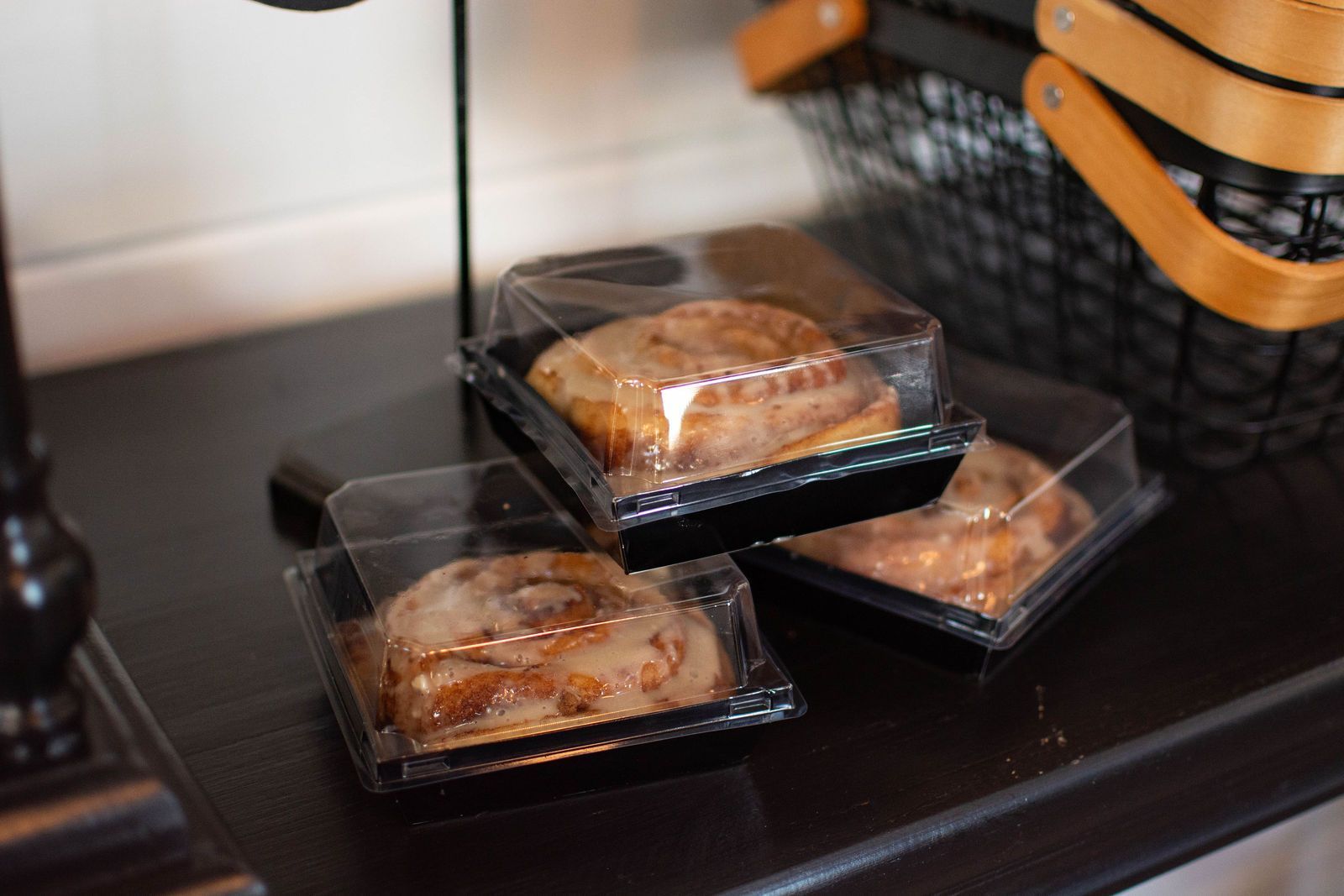 Three cinnamon rolls in clear plastic containers on a dark wooden table next to a black basket.