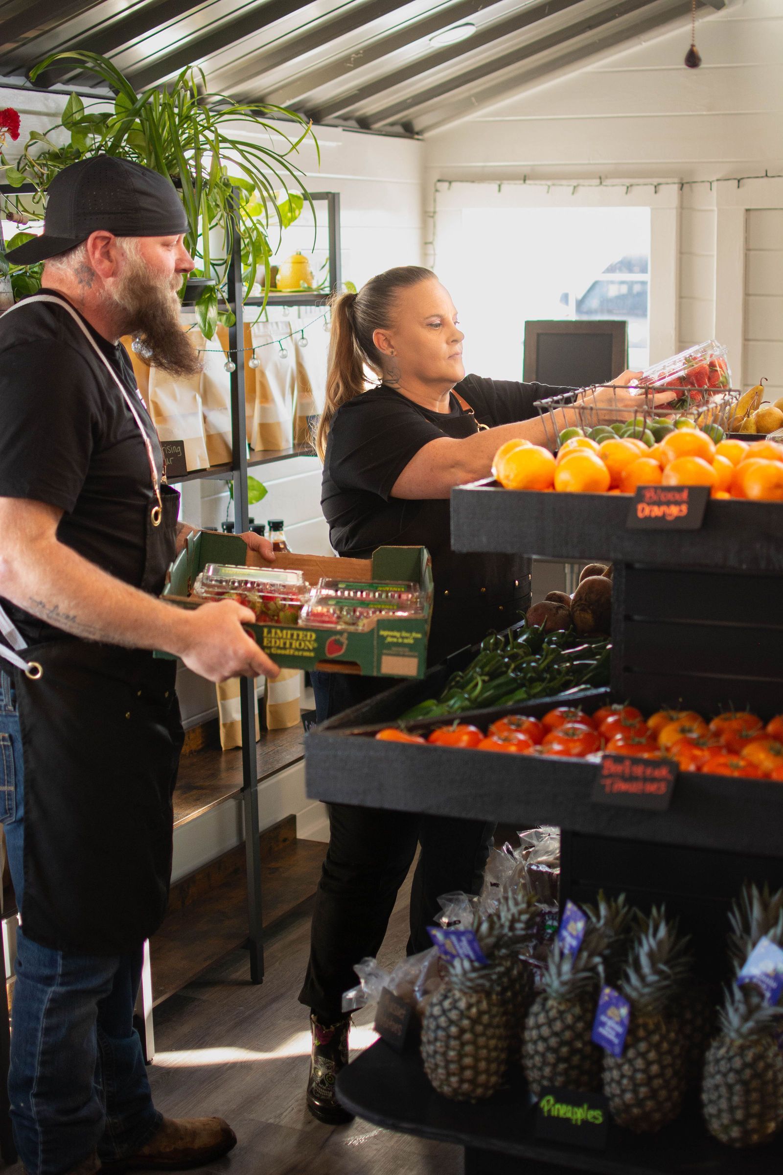 Two people stocking produce in a farm stand. Man holds a carton, woman arranges fruit on a shelf. Bright interior.