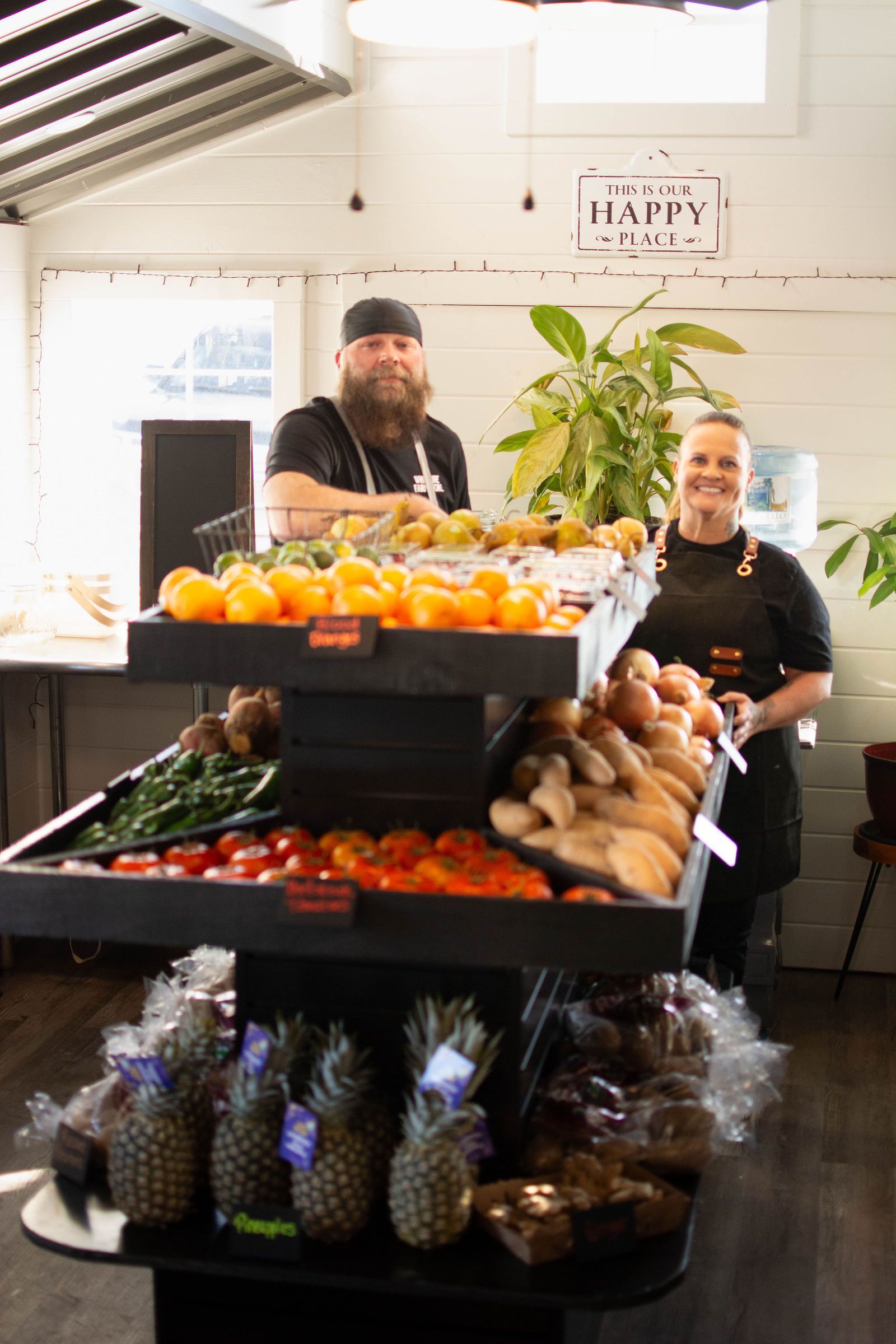 Two people behind a tiered display of produce. Displaying pineapples, tomatoes, and greens.
