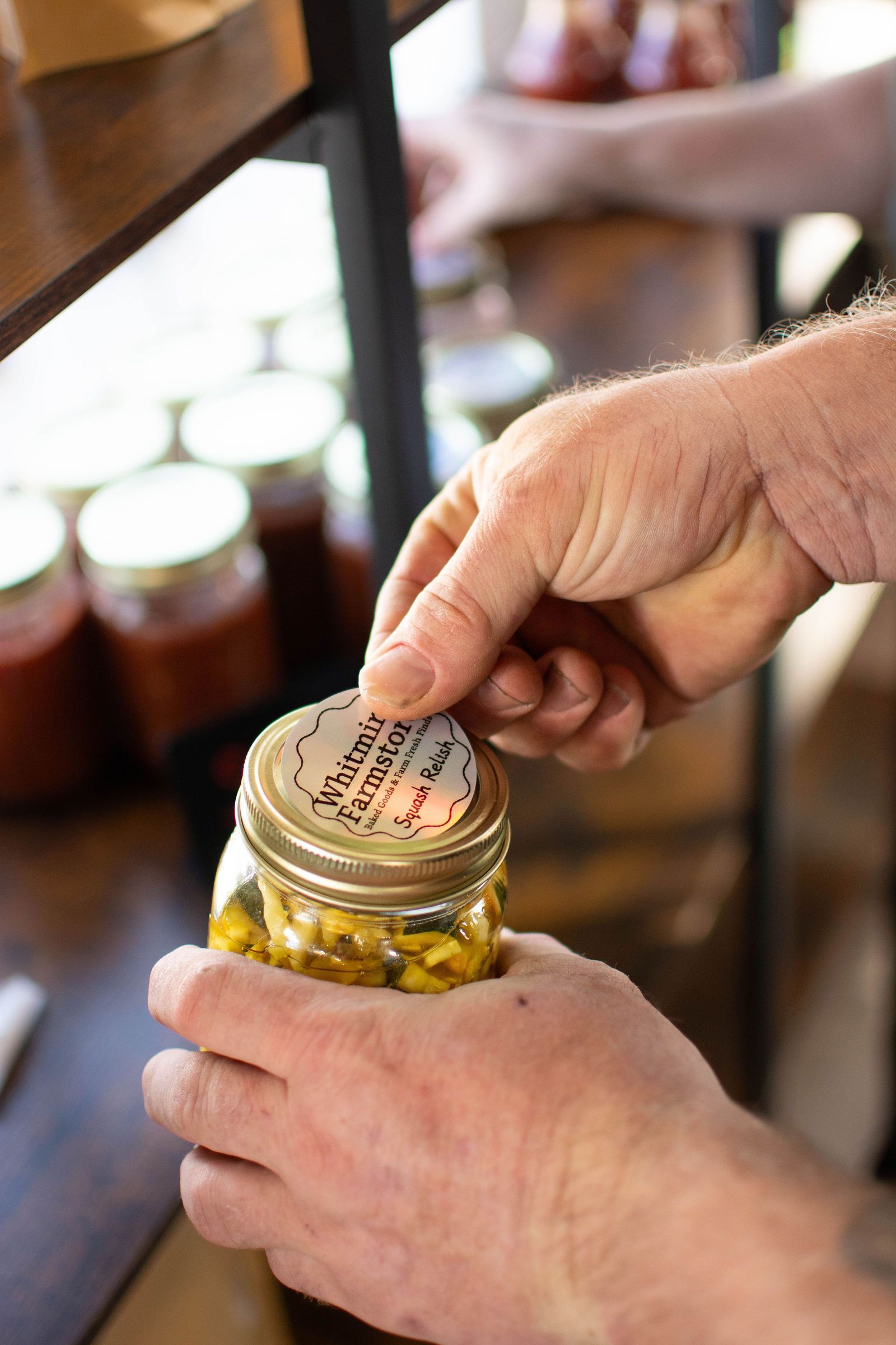 Hands peeling a sticker off a jar lid. Jars of food are on a wooden shelf in the background.