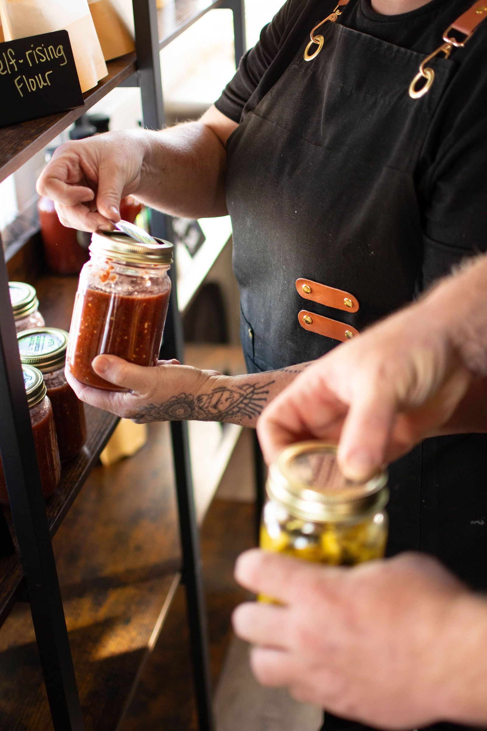 Person holding jars of preserved food on a shelf, likely in a market.