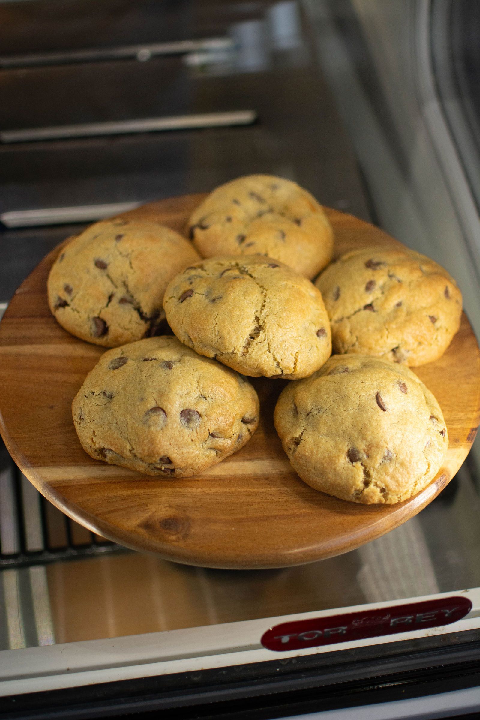 Six chocolate chip cookies on a wooden plate, inside a glass display.