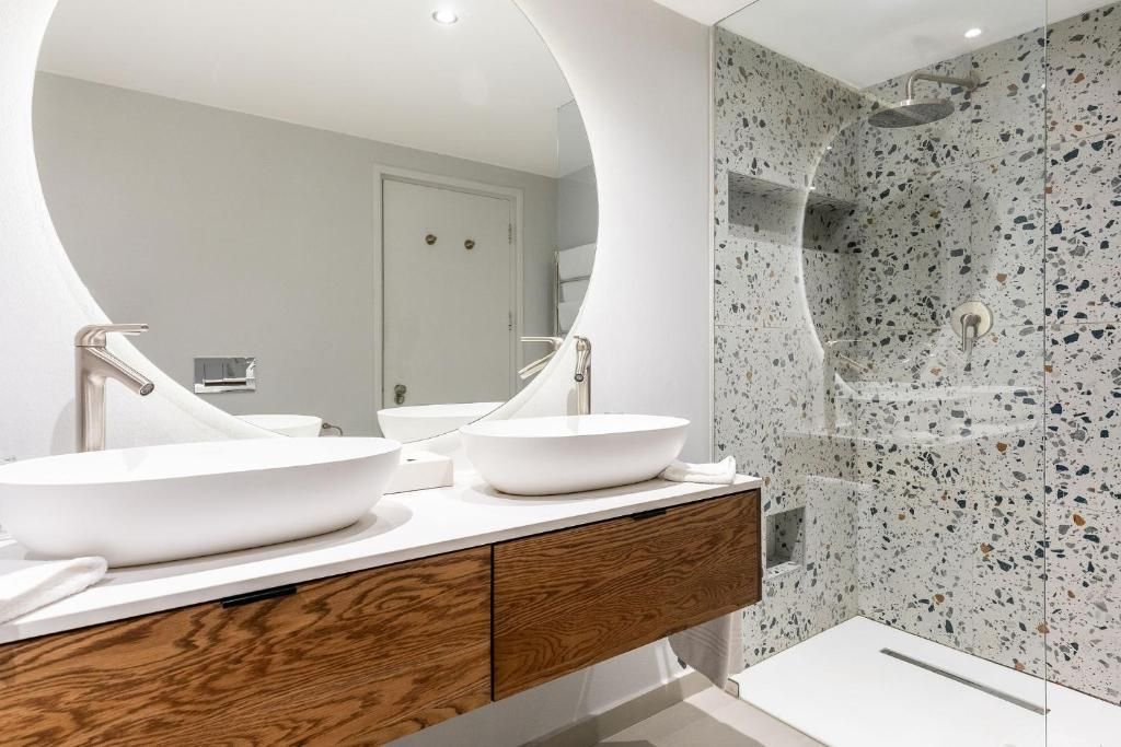 Modern bathroom with double sinks, large round mirror, and terrazzo-tiled shower.