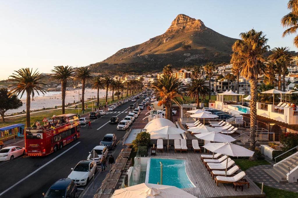 Roadside view of Lion's Head mountain in Cape Town. Red bus, palm trees, pool with white umbrellas, beach, and cars.