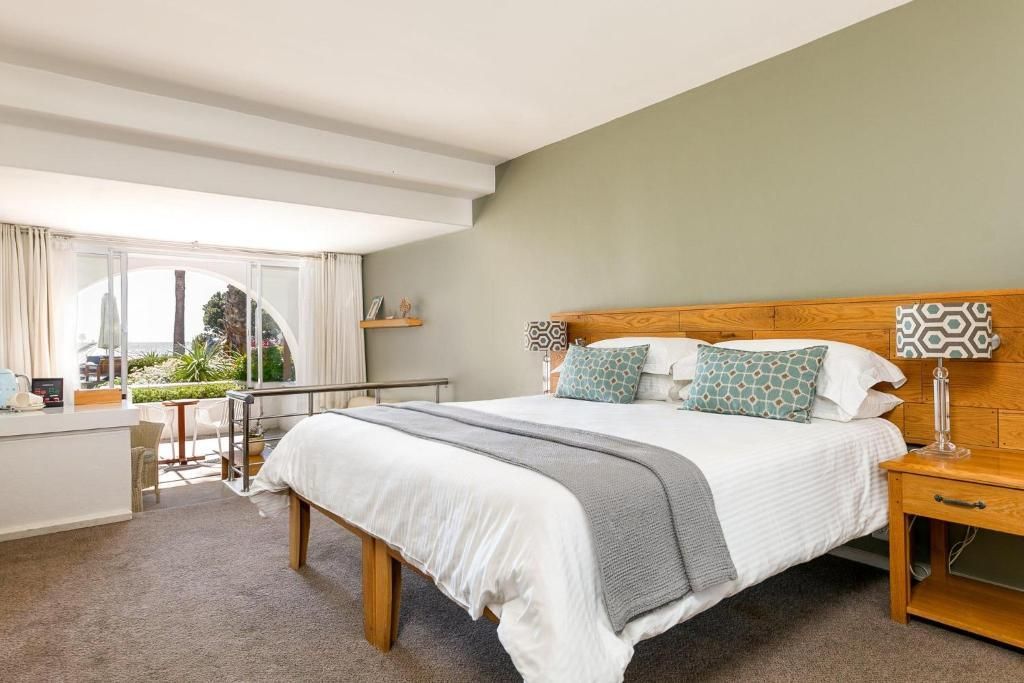 Bedroom with a large bed, nightstand, desk, and balcony overlooking the ocean; neutral color palette.