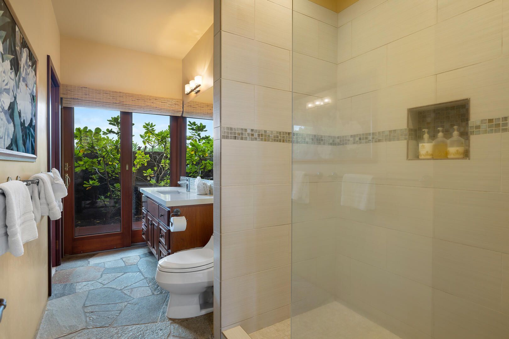 Bathroom with shower, toilet, and vanity, glass shower door, window, and outdoor foliage.