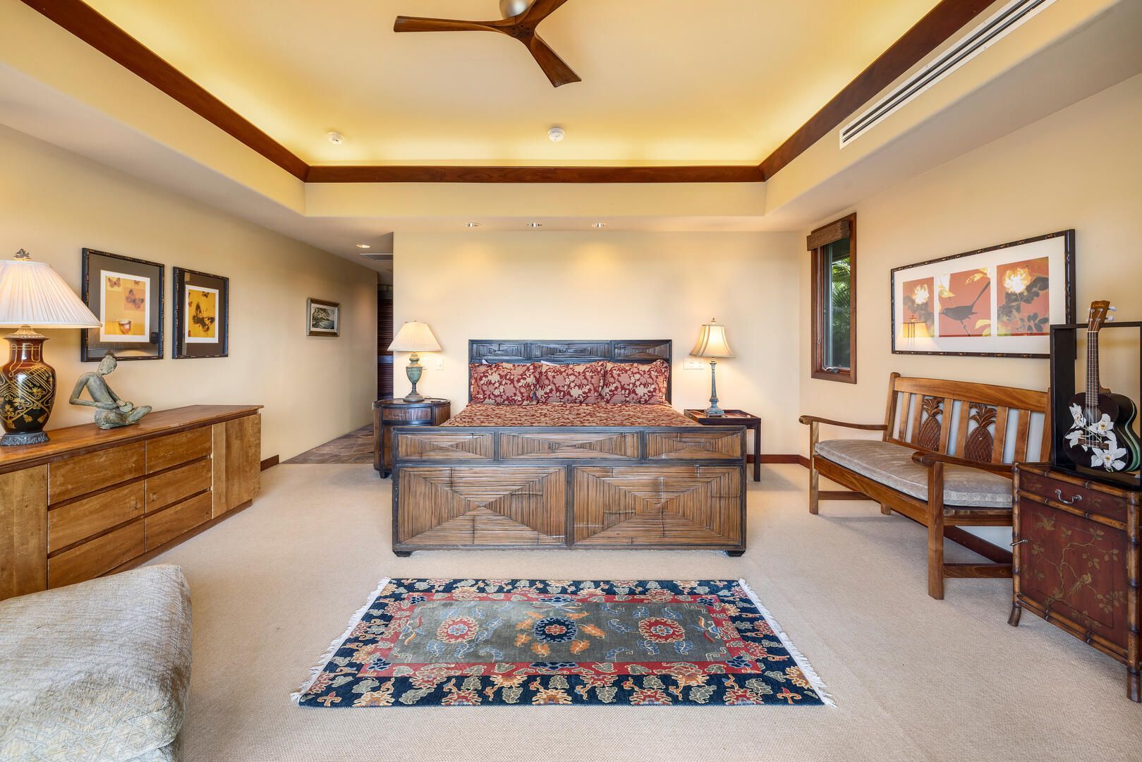 Spacious bedroom with a large wooden bed, rug, dresser, bench, art, and ceiling fan.