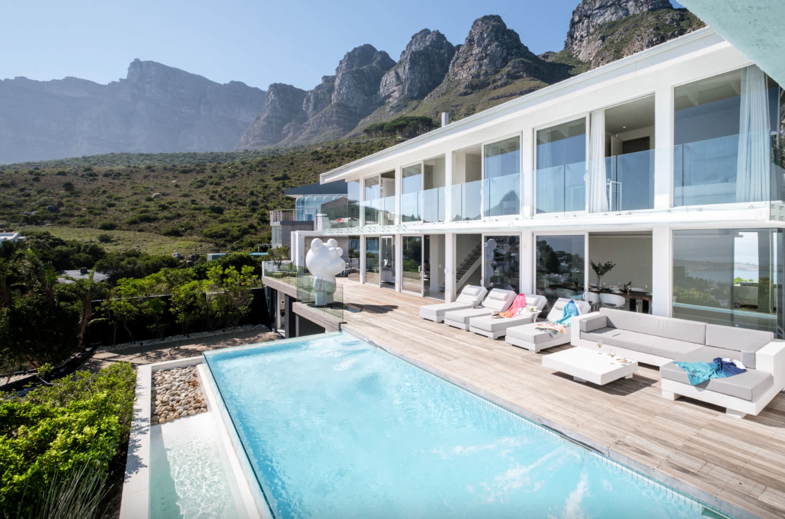 Luxurious white house in Cape Town with pool and deck, overlooking mountain range and ocean.