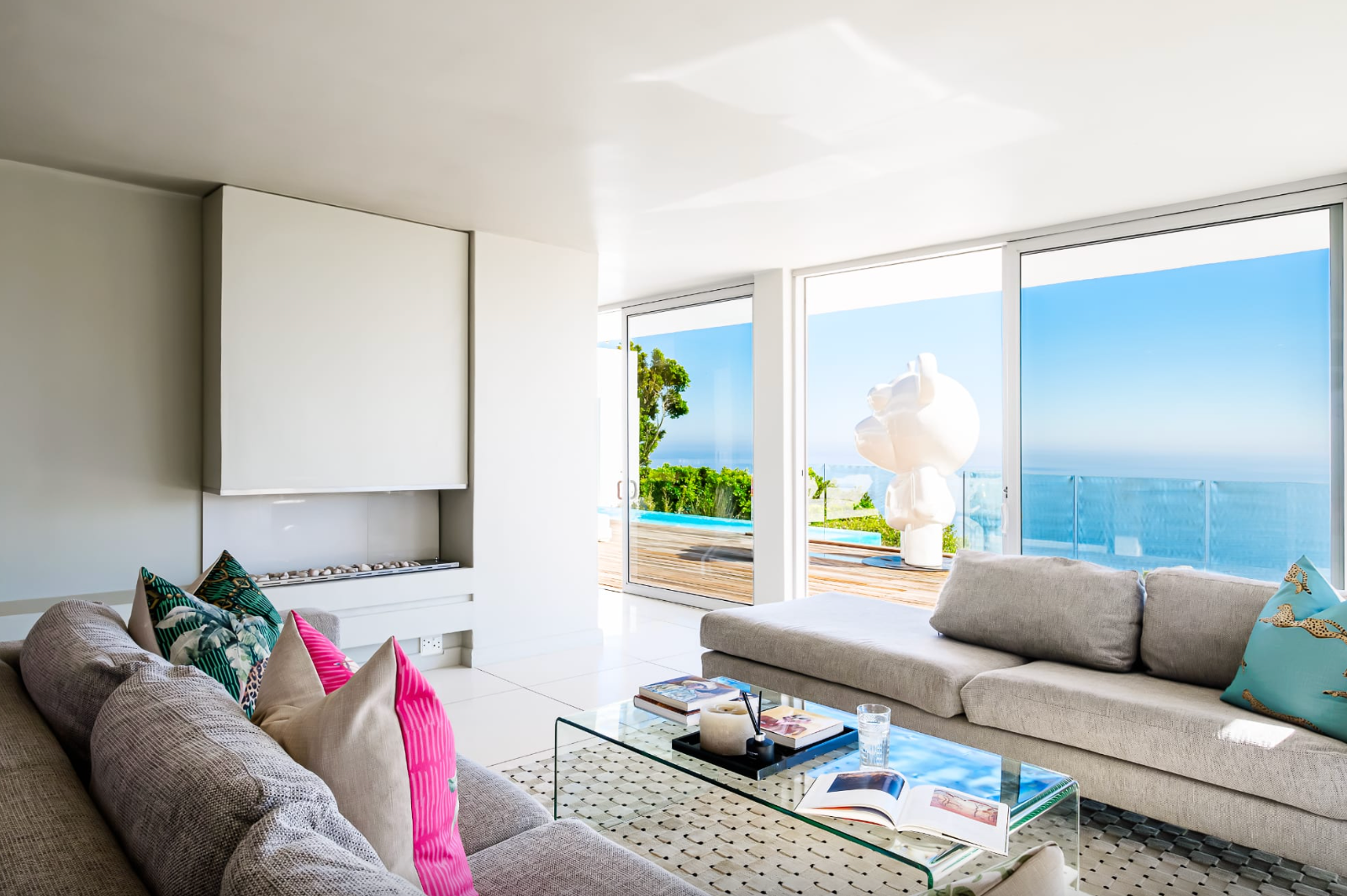 Bright modern living room with ocean view, gray sofa, glass coffee table, colorful pillows.