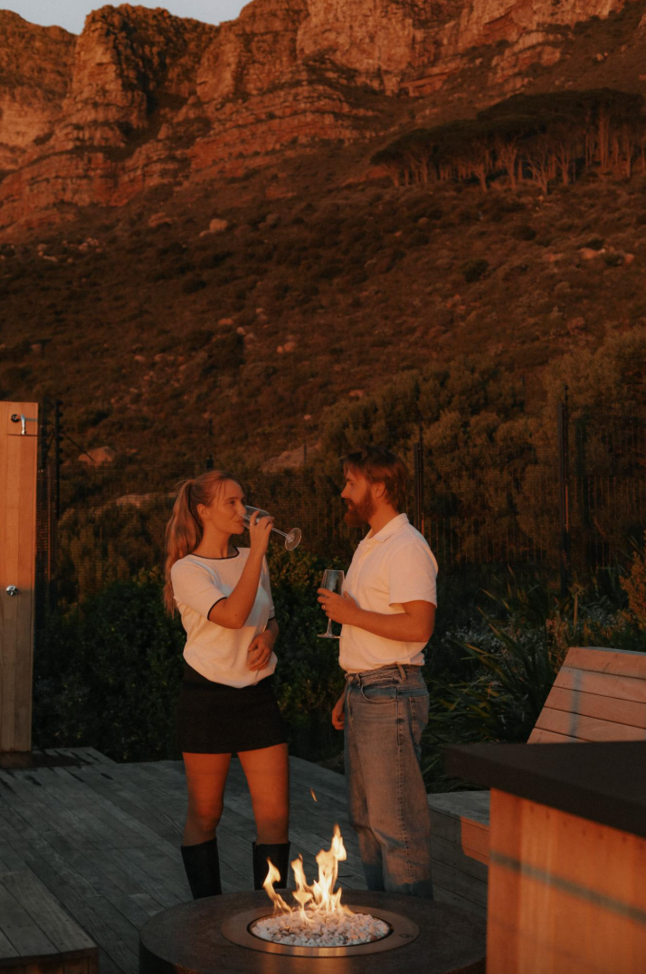 Couple enjoying wine by a fire pit, with mountain backdrop at sunset.