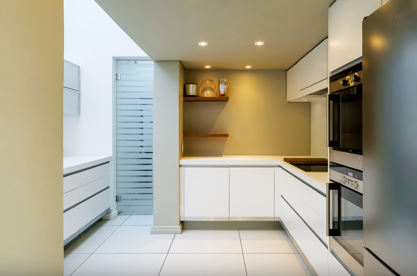 Modern white kitchen with built-in appliances, recessed lighting, and a frosted glass door.