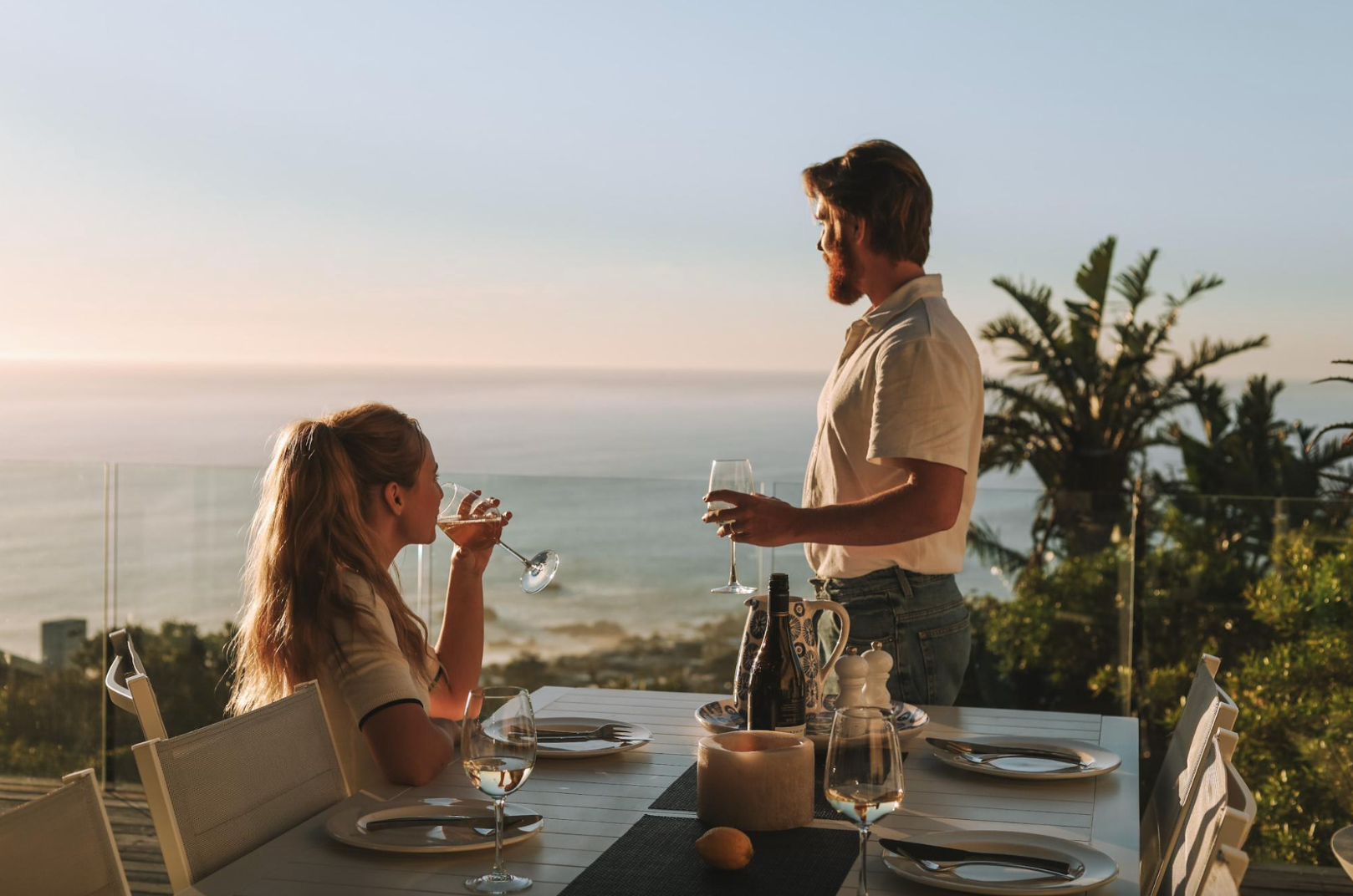 Couple enjoying wine on a patio overlooking the ocean. Sunny day, table set for dinner.