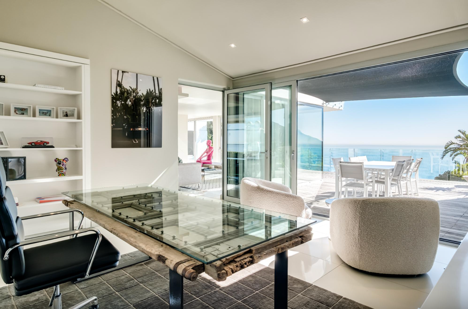 Office with a glass desk, white bookshelf, and ocean view through open glass doors.