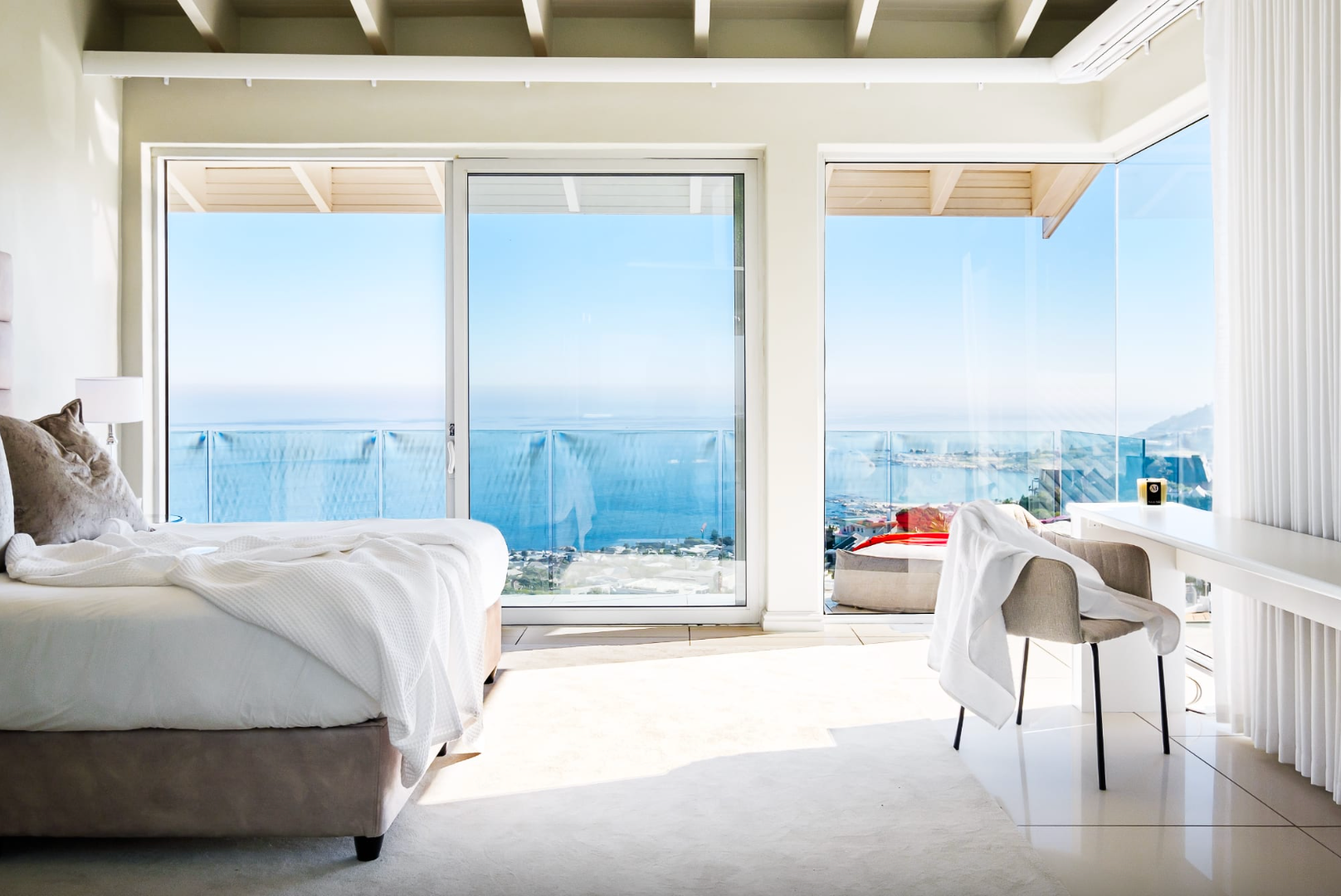 Bedroom with ocean view; bed, desk, and sliding glass doors. Bright, airy space with blue water.