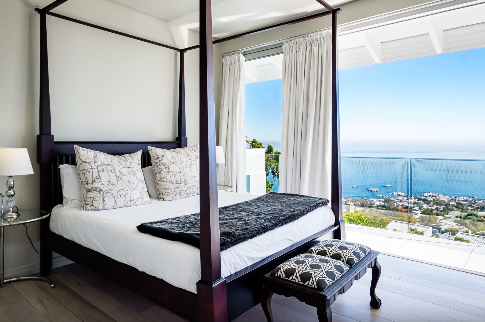 Bedroom with dark wood canopy bed, white bedding, bench, and ocean view.