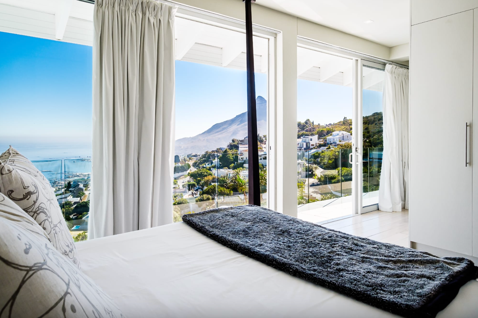 Bedroom with a bed, pillows, and a window overlooking a cityscape and mountain against a blue sky.