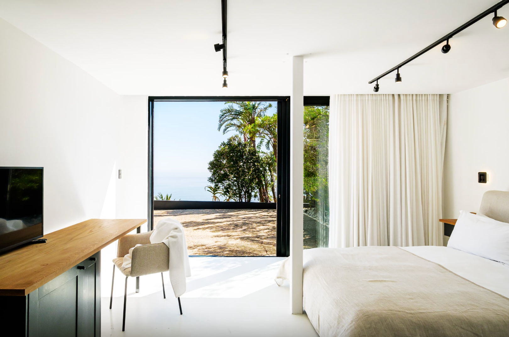 Bright bedroom with a view of trees and water. Modern decor with a bed, desk, chair, and large window.